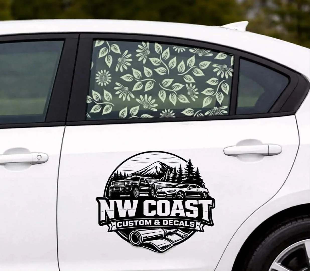 A white car with a NW Coast Custom & Decals sticker on the door, featuring mountain, forest, and road illustrations. The window has a floral privacy decal with white leaves and flowers.