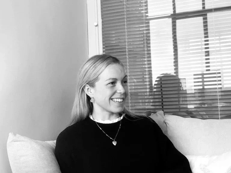 A woman sitting on a couch, smiling, with blinds and a window in the background.