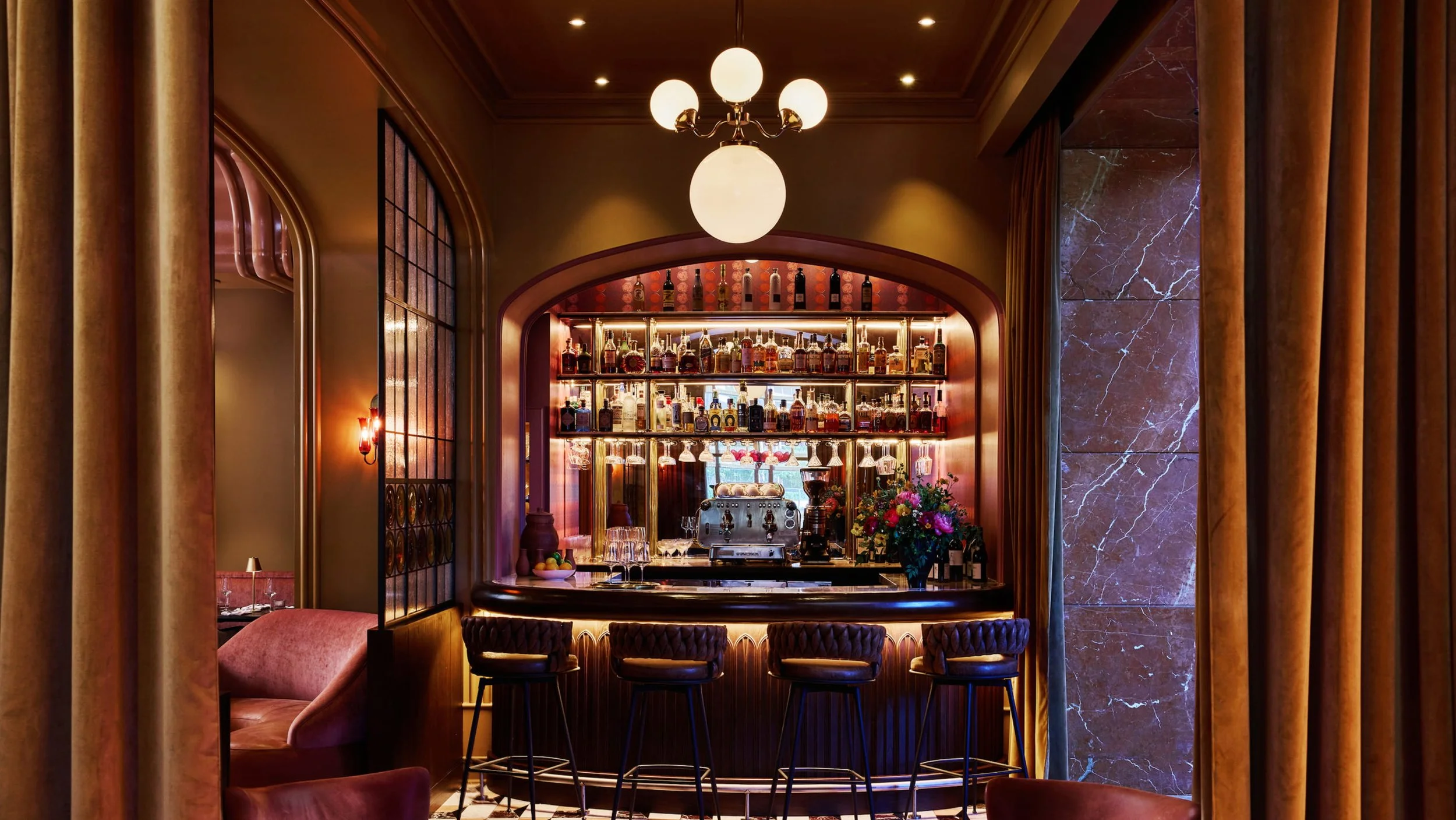 A luxurious bar with a marble countertop, equipped with four bar stools, surrounded by warm lighting, purple curtains, and a background with a collection of liquor bottles and a coffee machine.