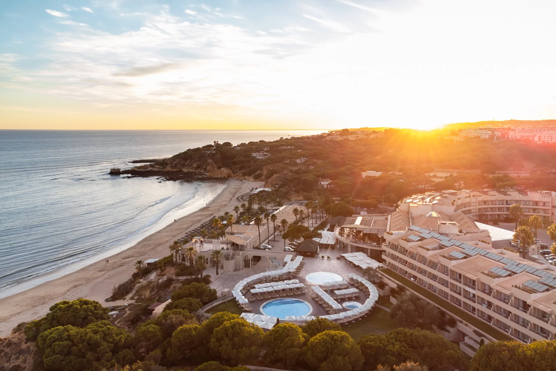 Aerial view of a beach resort at sunset, with a sandy beach, a swimming pool, luxury buildings, and lush greenery, overlooking the ocean.