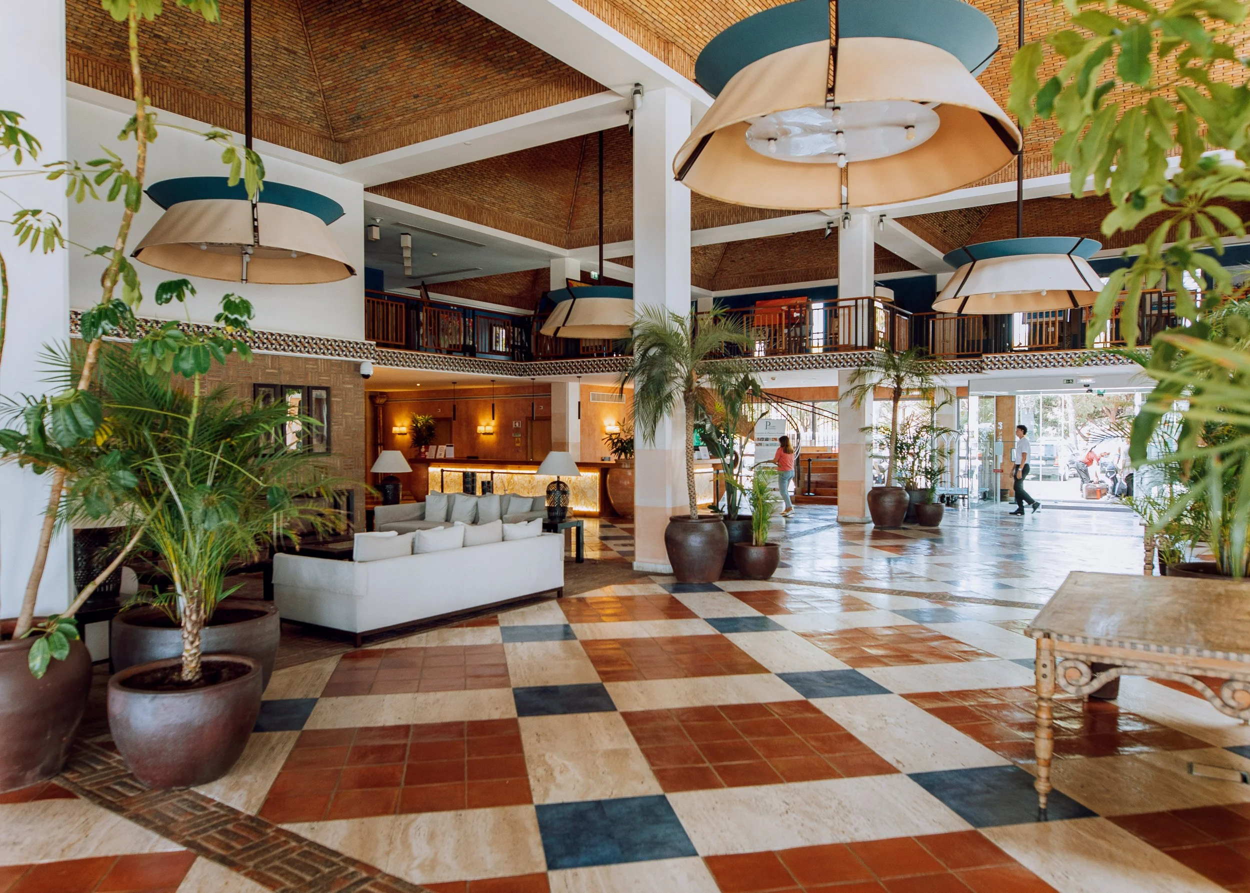 Interior of a hotel lobby with large pendant lights, potted plants, a white sofa, wooden and tiled flooring, and a reception desk in the background. There are people walking and seated in the lobby.