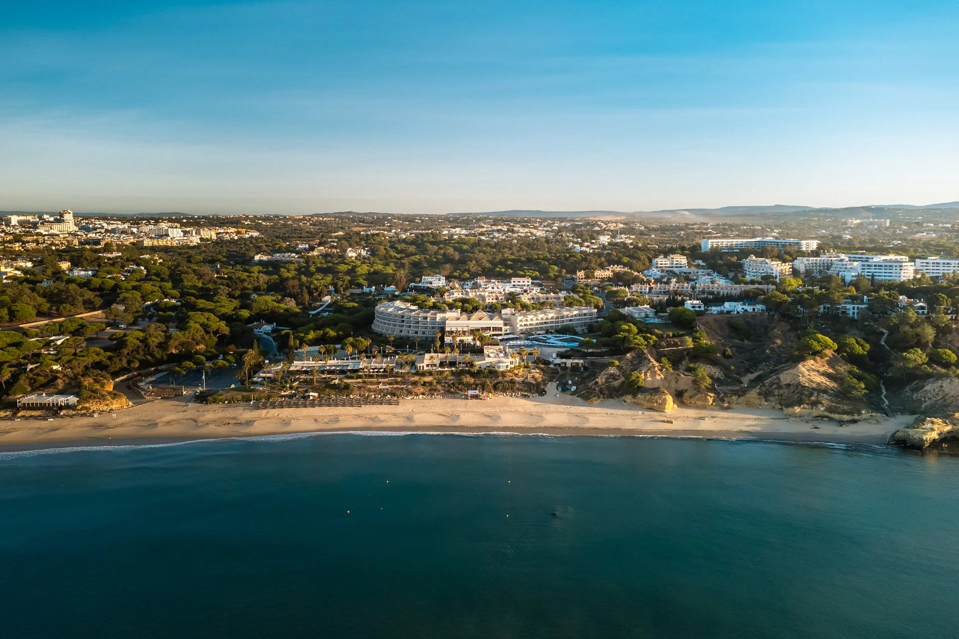 Aerial view of a coastal city with a sandy beach, turquoise water, and residential buildings surrounded by green trees.