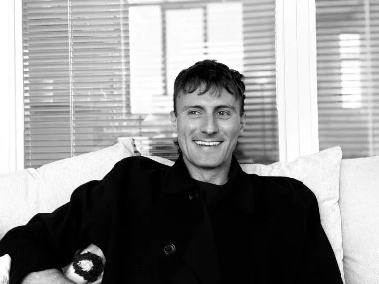 A young man with short, tousled hair is smiling and sitting on a light-colored sofa in front of a window with blinds, wearing a dark jacket.