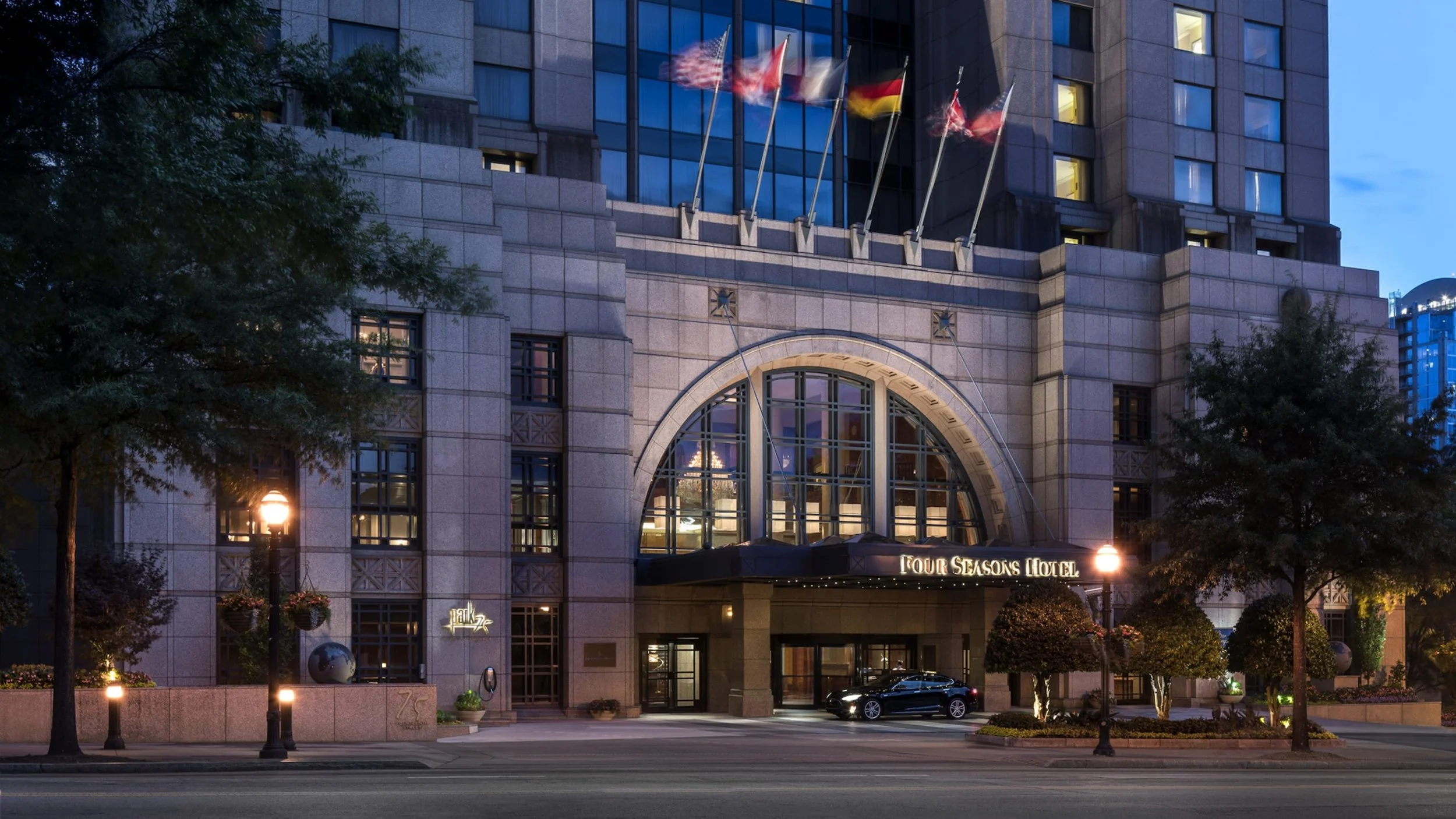 Night view of the Four Seasons Hotel's entrance with lit street lamps, trees, and flags, showing the hotel building facade and car parked outside.