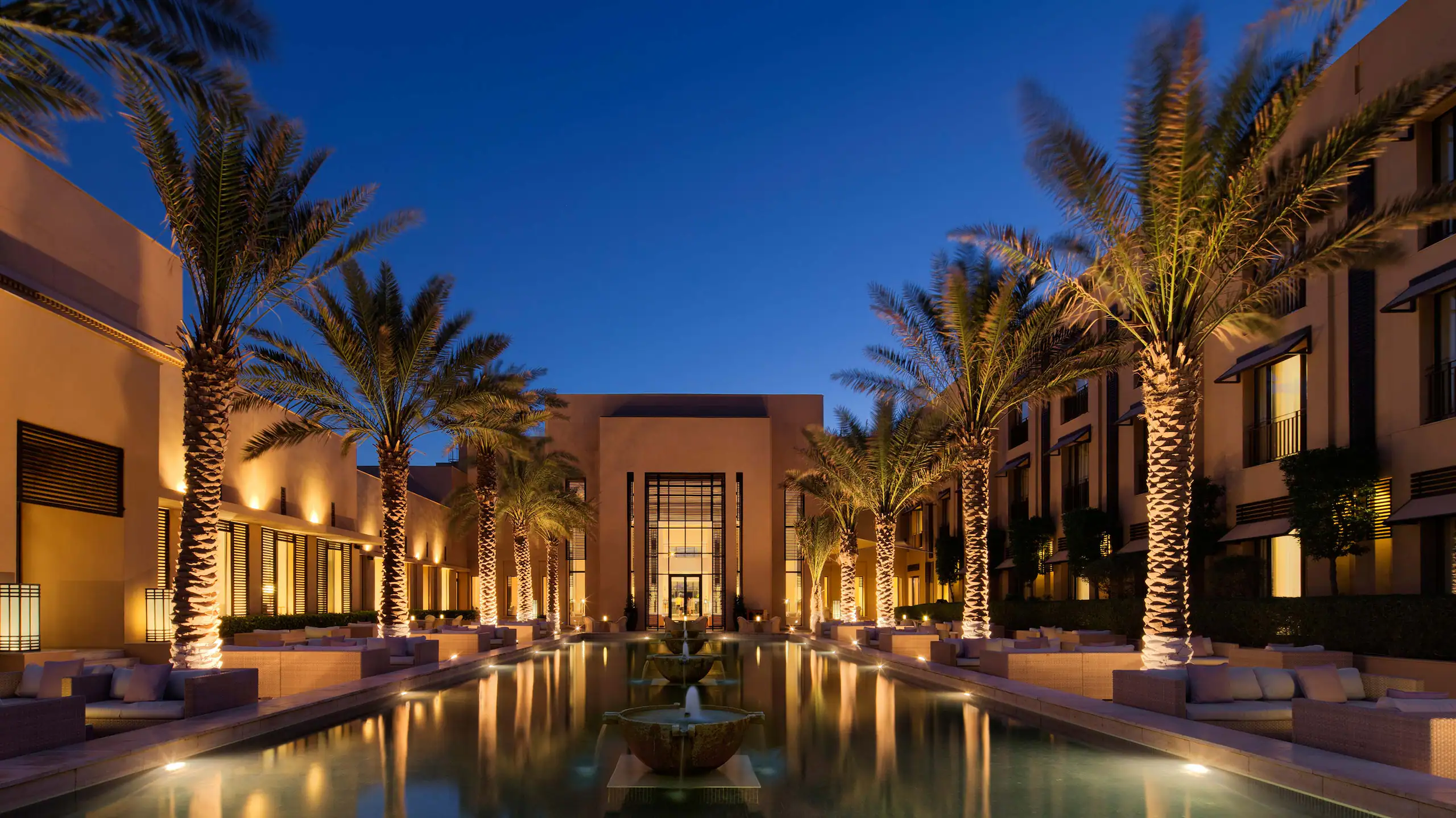 Luxury hotel courtyard at night with tall palm trees, water fountain, and illuminated seating areas, modern architecture, clear evening sky.