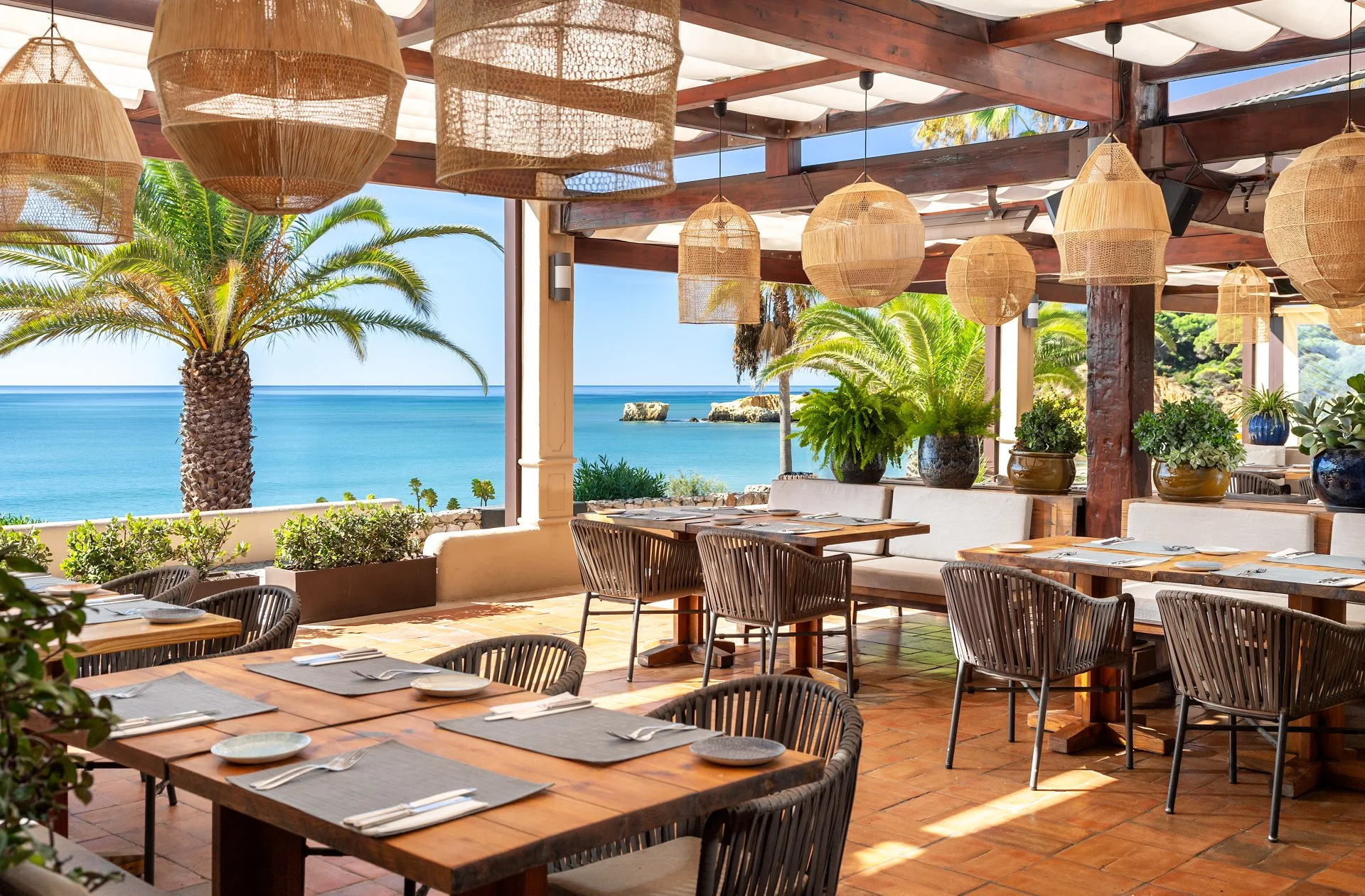 Indoor restaurant with wooden tables and chairs, overlooking a patio with palm trees and a view of the ocean