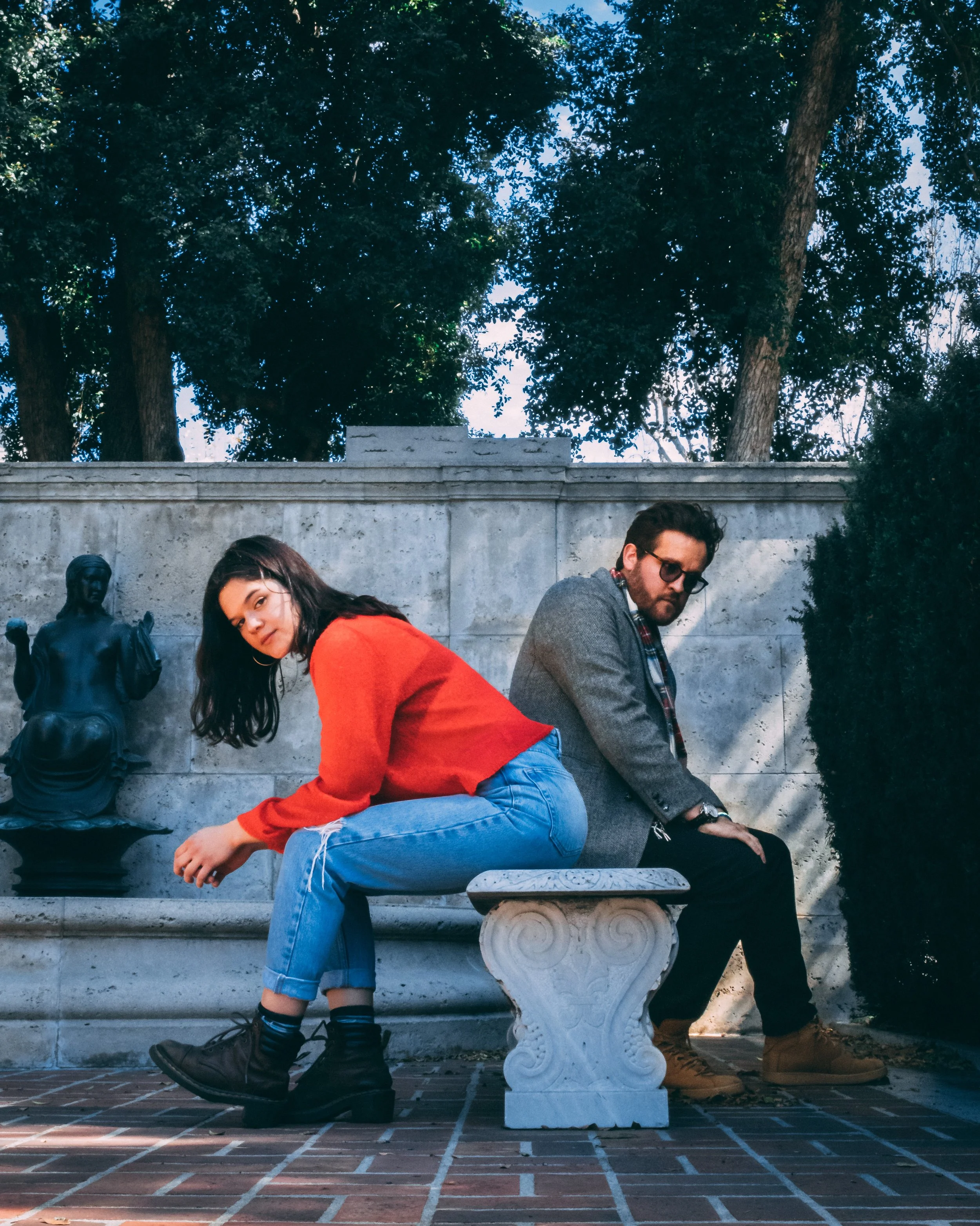 Two people sitting on a concrete bench, with a woman in an orange sweater and blue jeans leaning forward and a man in a gray blazer and glasses sitting beside her, both in an outdoor park area with trees in the background.