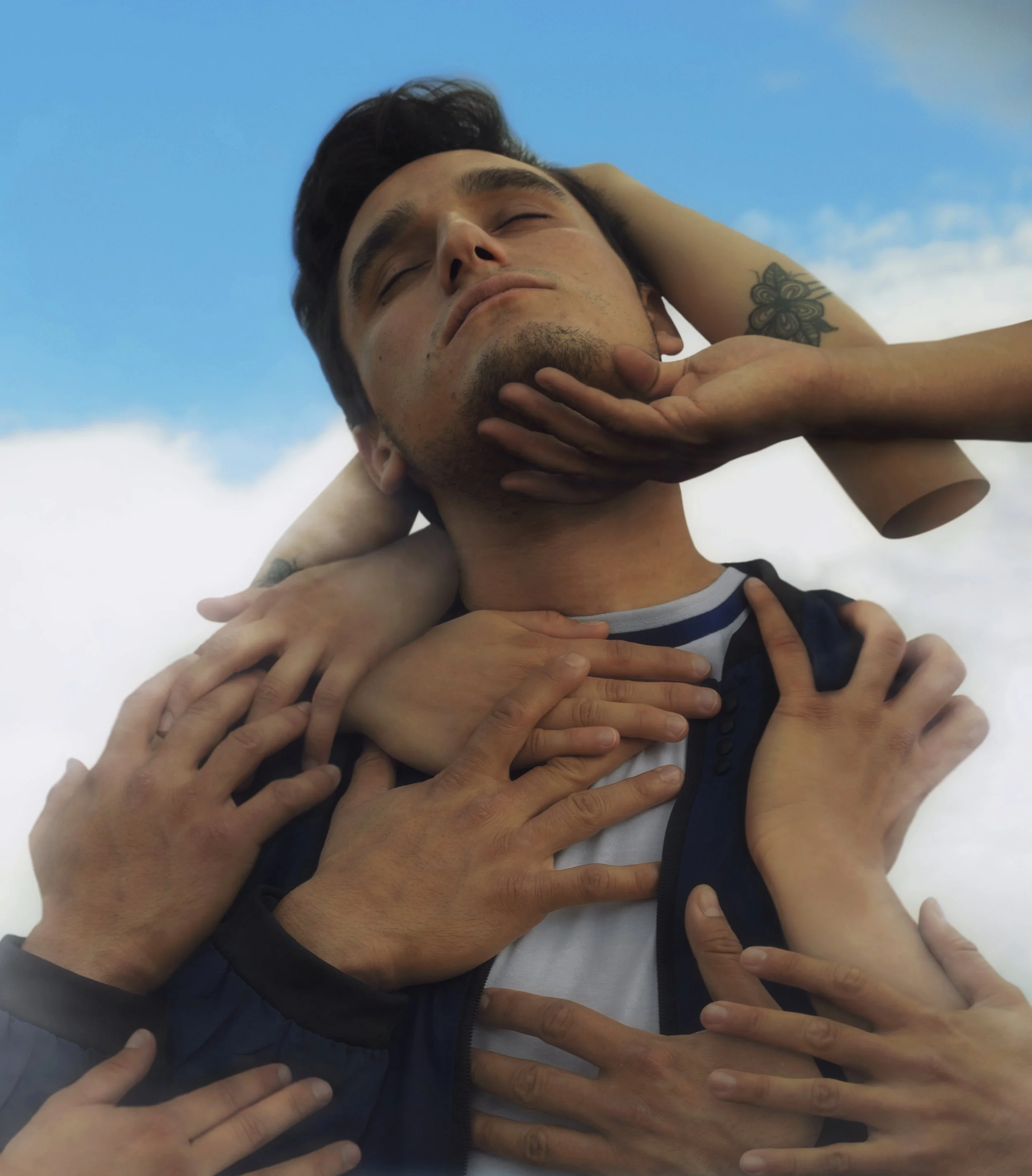 A man with dark hair and a tattoo on his arm is lying with his eyes closed, surrounded by multiple hands touching his head, neck, and shoulders against a blue sky background.