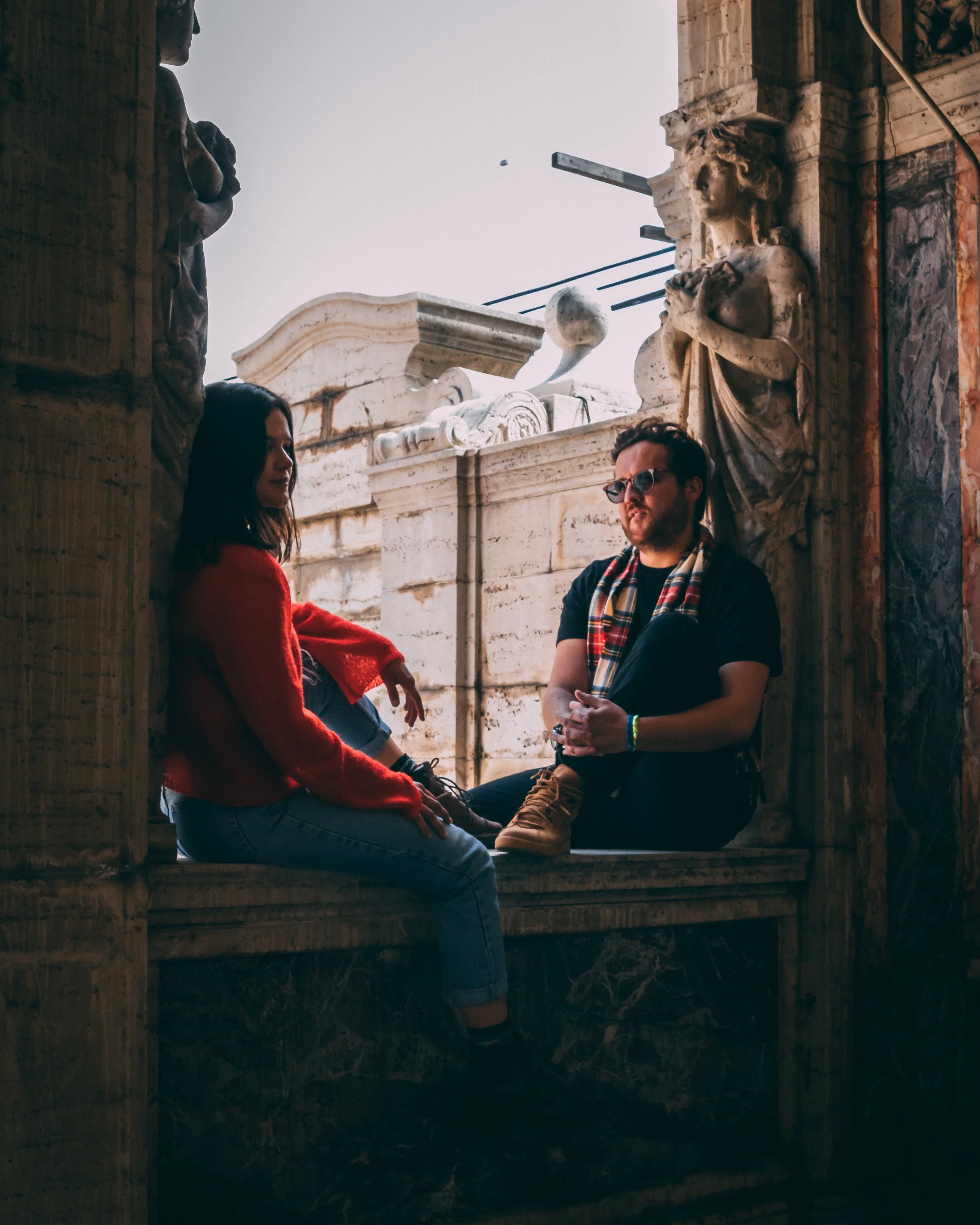 Two people sitting on a ledge in a historic or classical architectural setting with ornate stone sculptures and columns. A woman in a red sweater and a man with glasses and a plaid scarf are engaged in conversation.