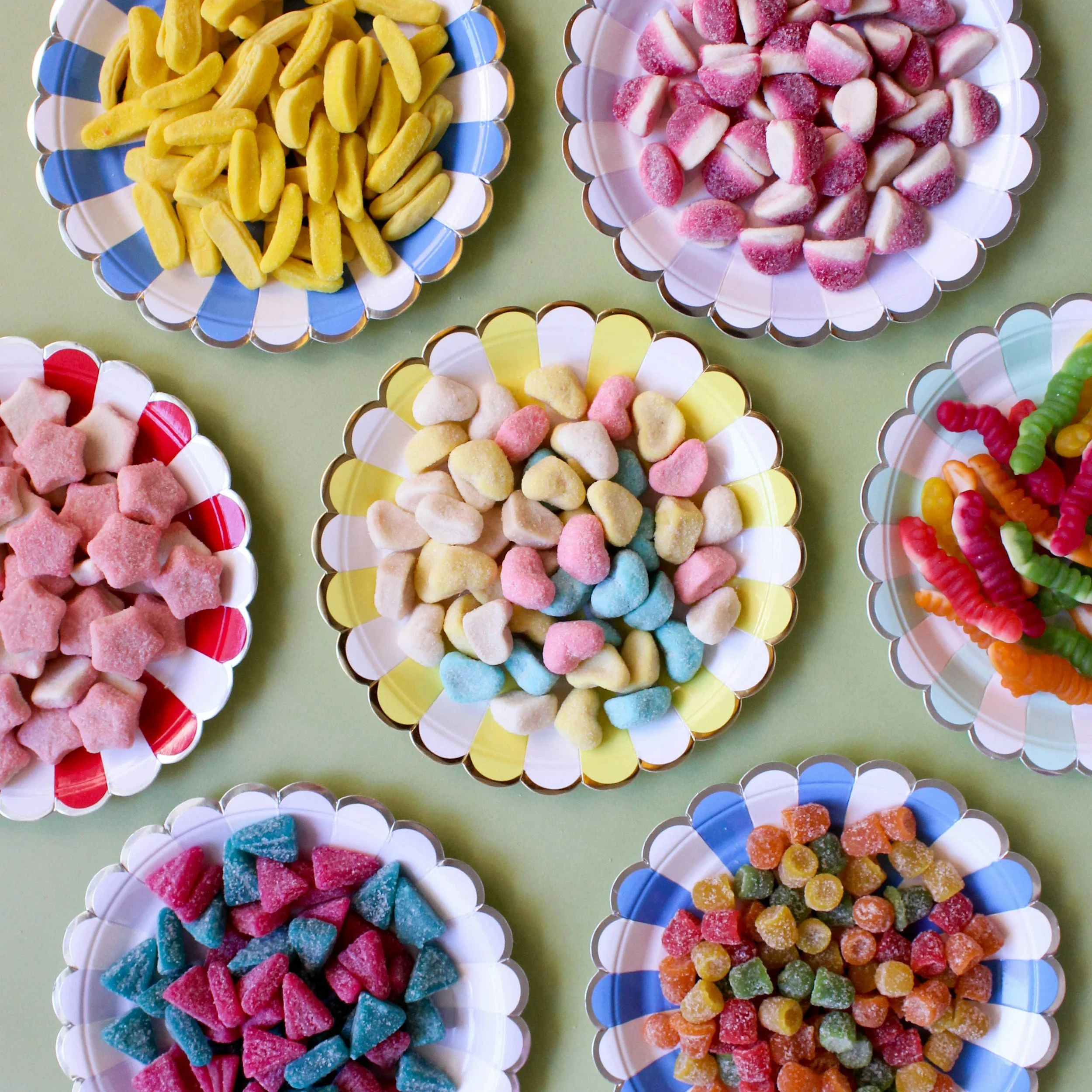 Colorful bowls filled with various types of candy, including gummy worms, star-shaped gummies, and multi-colored sugar-coated candies, arranged on a light green surface.