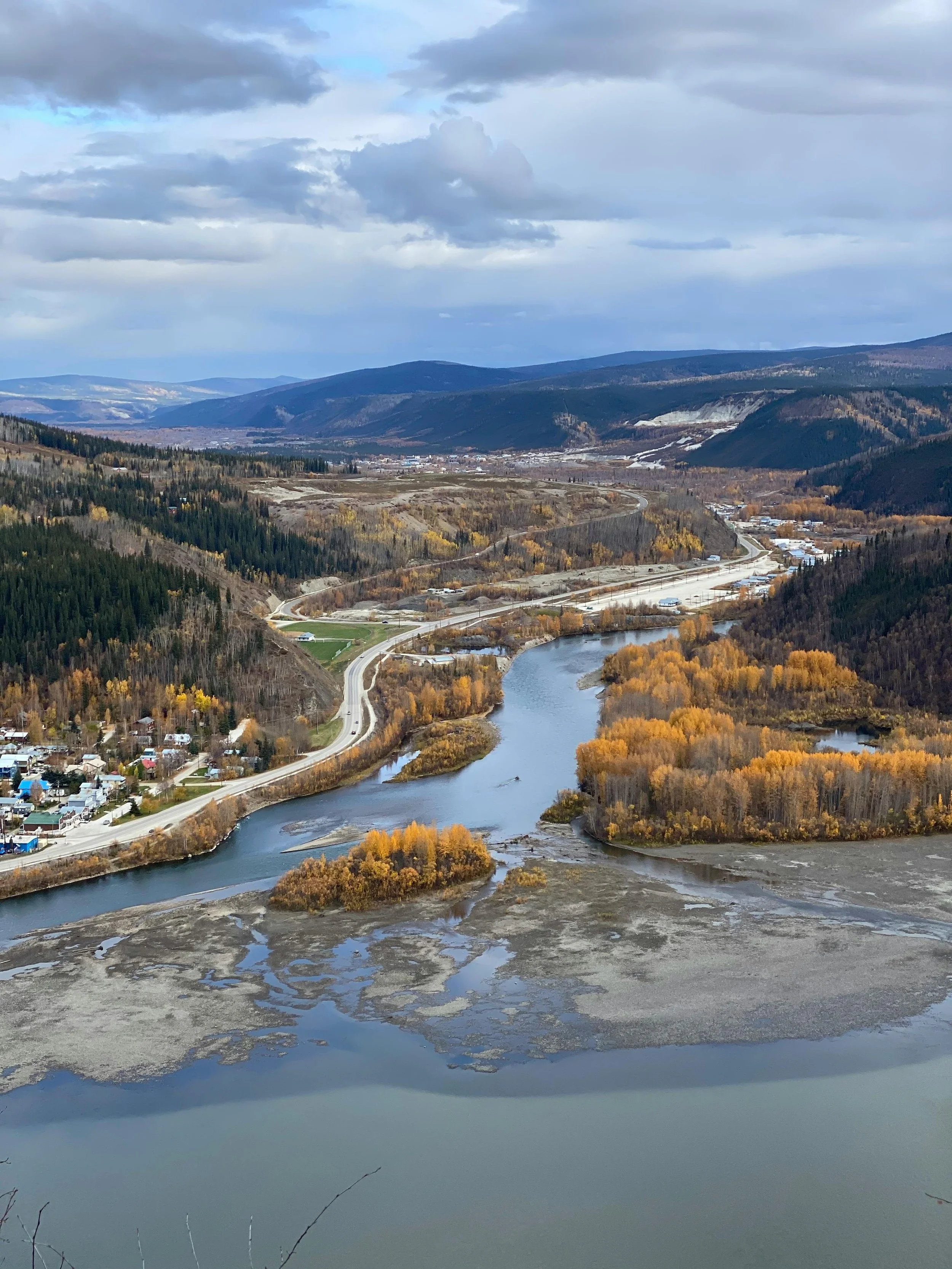 Aerial view of a winding river through a valley with yellow and orange autumn trees, surrounded by forested hills and mountains, under a cloudy sky.