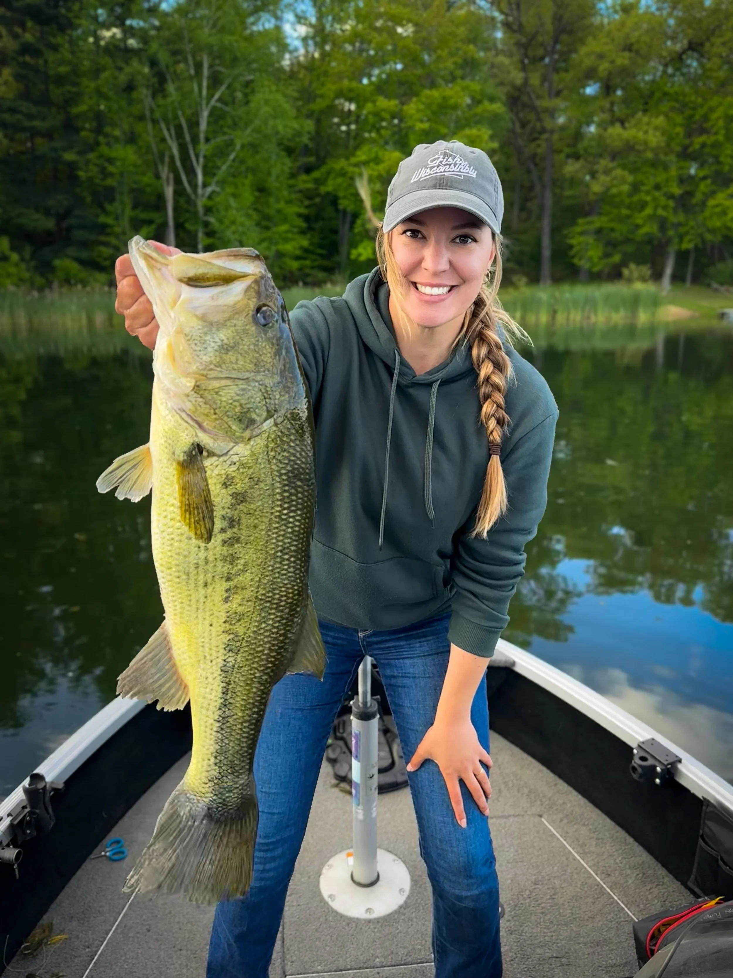 A woman with a braid, wearing a gray cap and a dark hoodie, is smiling and holding a large fish on a boat in a lake with green trees in the background.