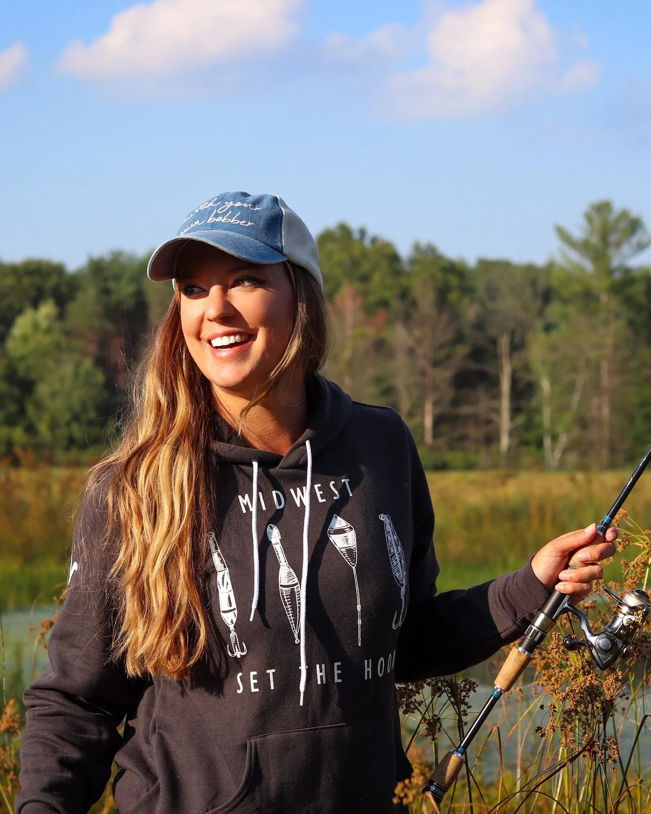 A woman smiling outdoors holding a fishing rod, wearing a hoodie and a baseball cap, with a lake and trees in the background.