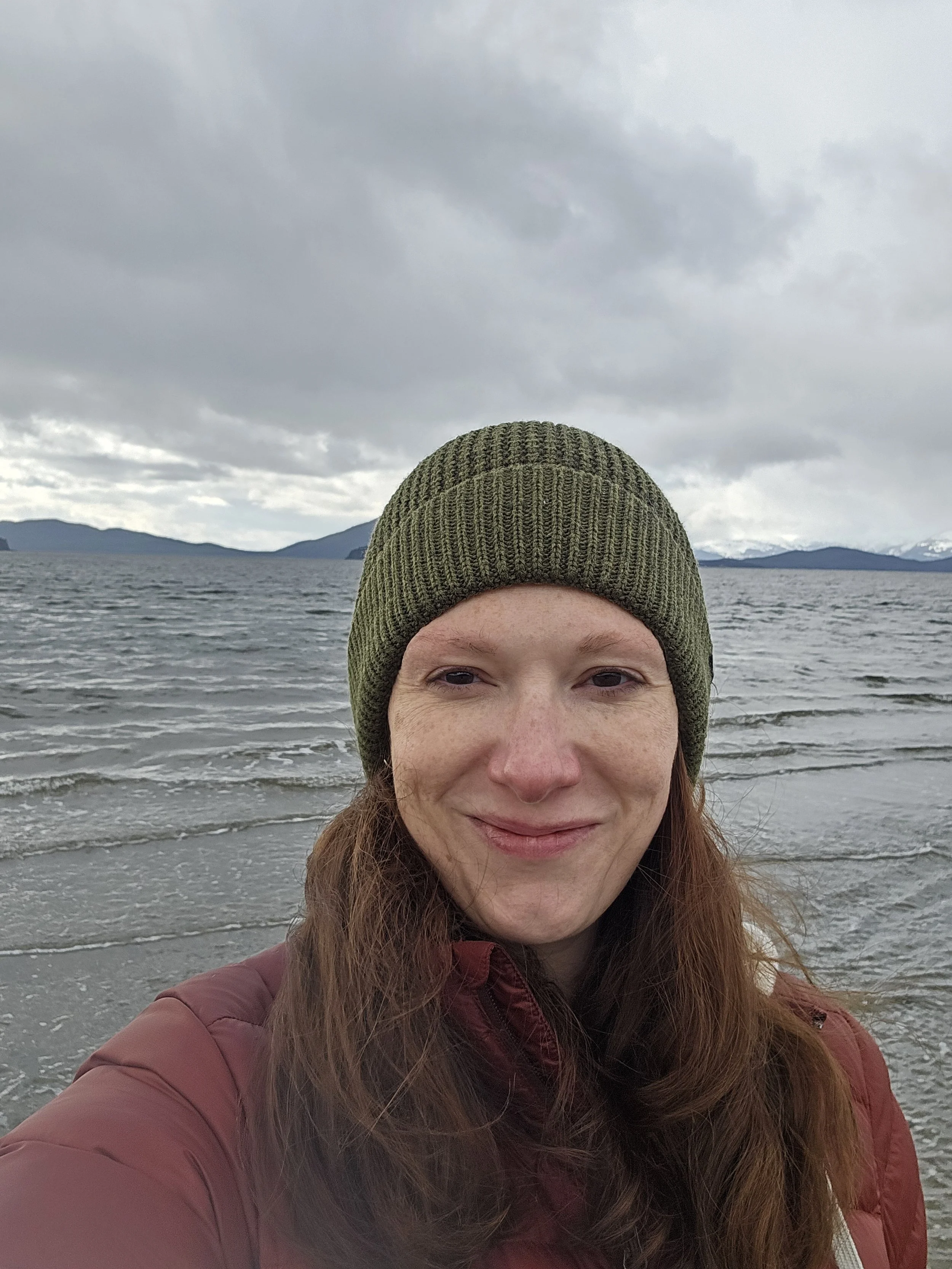 A woman with red hair wearing a green knit beanie and a maroon jacket, standing on a beach with waves, mountains, and cloudy sky in the background.