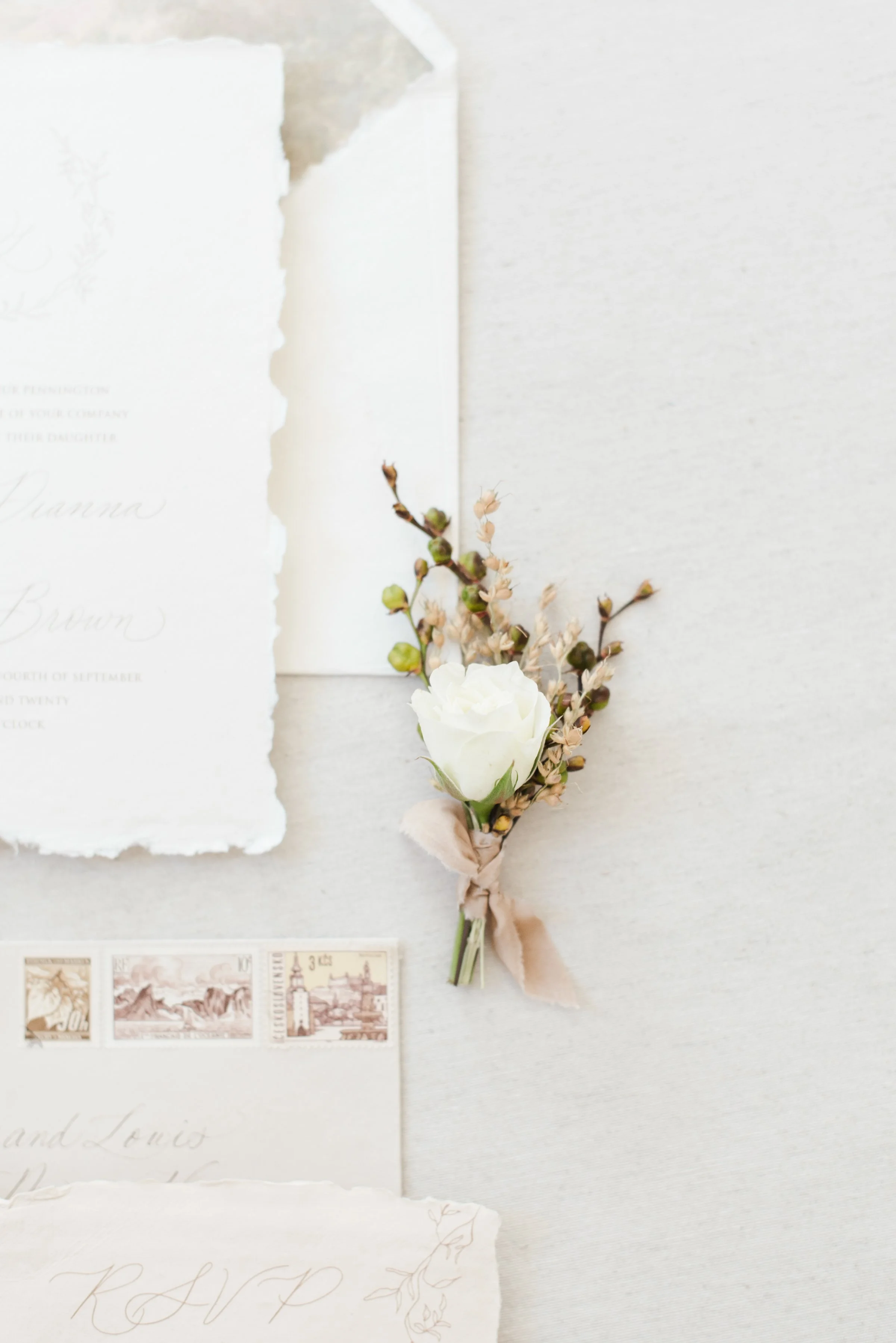 Wedding invitation suite on textured handmade paper with deckled edges, featuring a floral boutonniere and vintage-postage stamps.