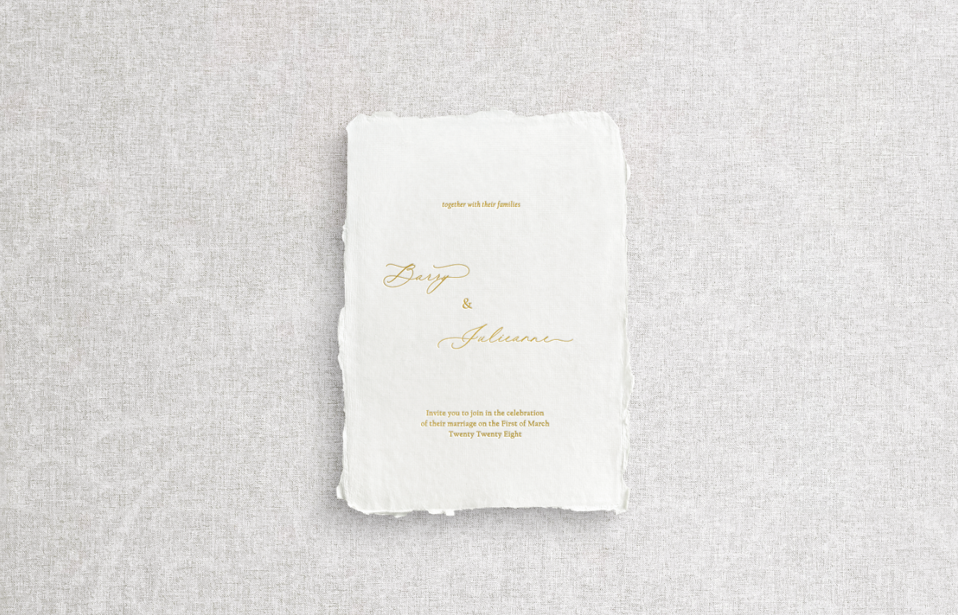 Main invitation on white handmade paper with gold foil letterpress on gray flat lay background.