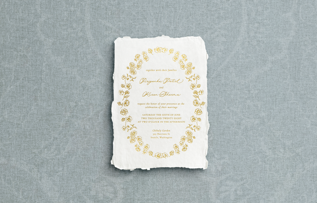 White invitation on handmade paper with floral botanical border encasing mixed typography of calligraphy printed using gold foil letter press on blue background.