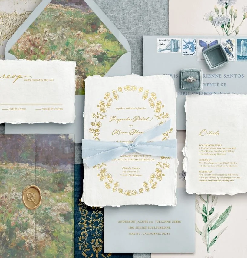 Wedding invitation mockup in a 'Timeless & Romantic' theme. Features deckled-edge handmade paper, gold foil lettering, and a floral wreath tied with dusty blue silk ribbon. Includes RSVP and details cards with vintage stamps and a gold wax seal.