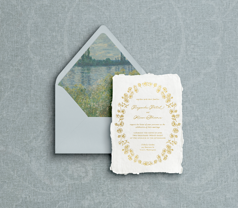 Blue envelope with watery scenic liner and white invitation on handmade paper with floral botanical border and mixed typography of calligraphy printed using gold foil letter press on blue background.