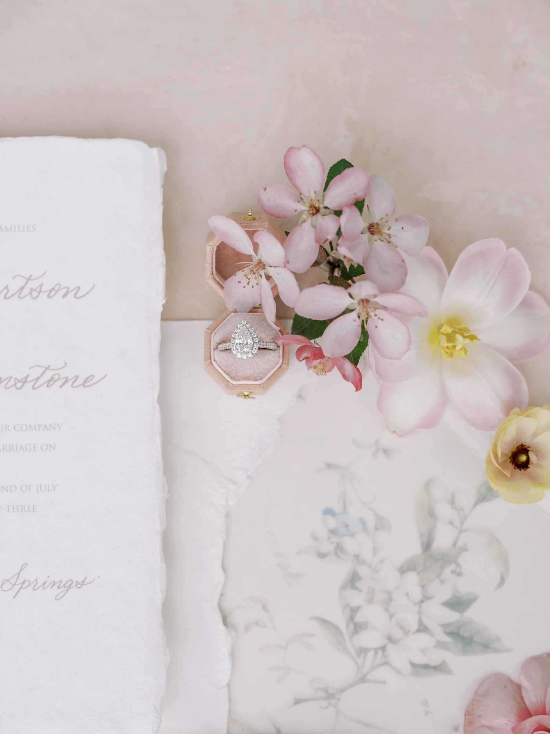 Close-up flatlay of a wedding invitation on handmade deckled-edge paper styled with pink spring flowers and an engagement ring in a velvet box.