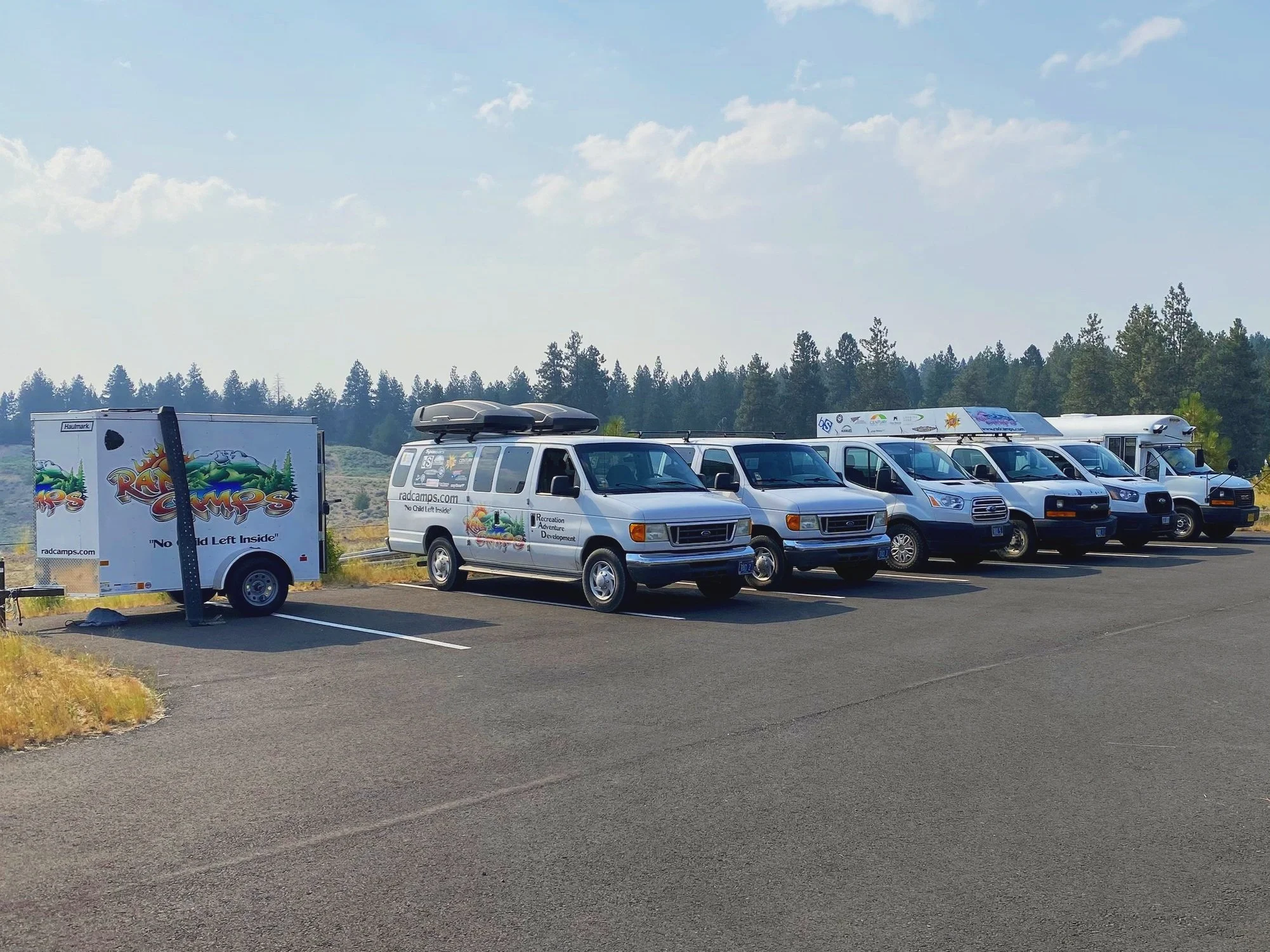 A row of recreational vehicles and a trailer parked in a lot with trees in the background under a partly cloudy sky.