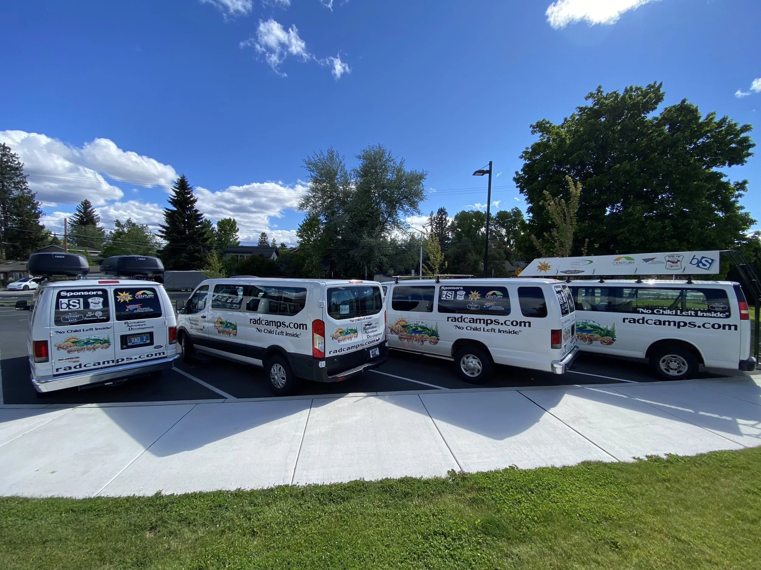 Four white vans parked in a row with promotional stickers and website URL 'radcamps.com', outdoors on a paved parking lot, with trees and a bright blue sky with clouds in the background.