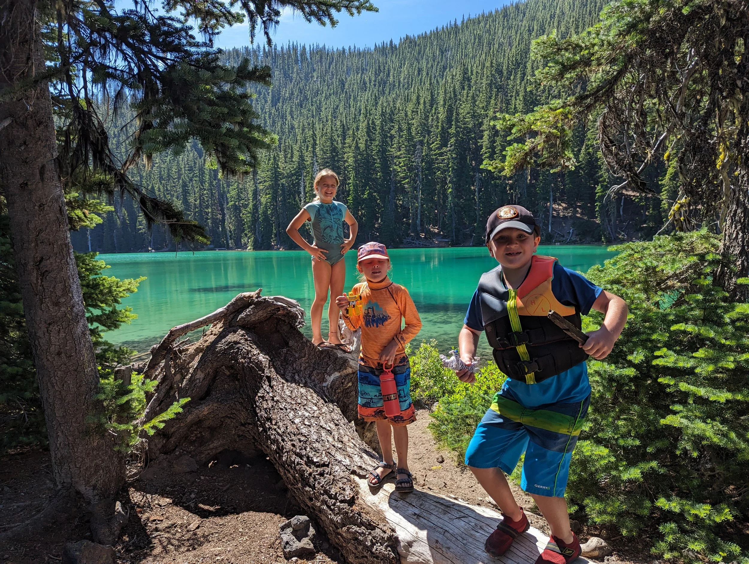Four children standing on a fallen tree by a lake with green water, surrounded by evergreen trees and mountain forest, during daytime.