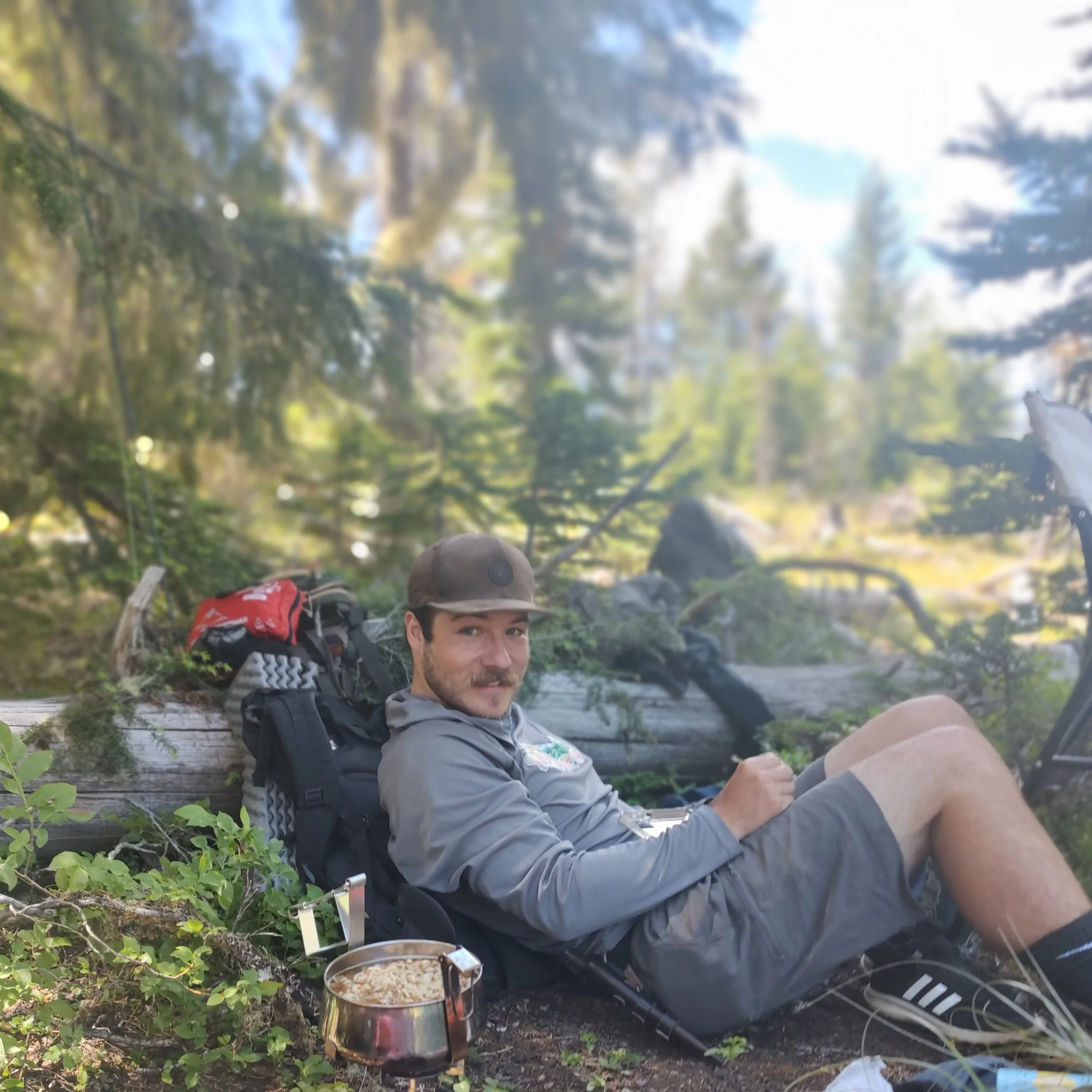 A man with a beard and mustache sitting in a forest, leaning against a log, with camping gear and a camping stove nearby, enjoying nature surrounded by trees and greenery.
