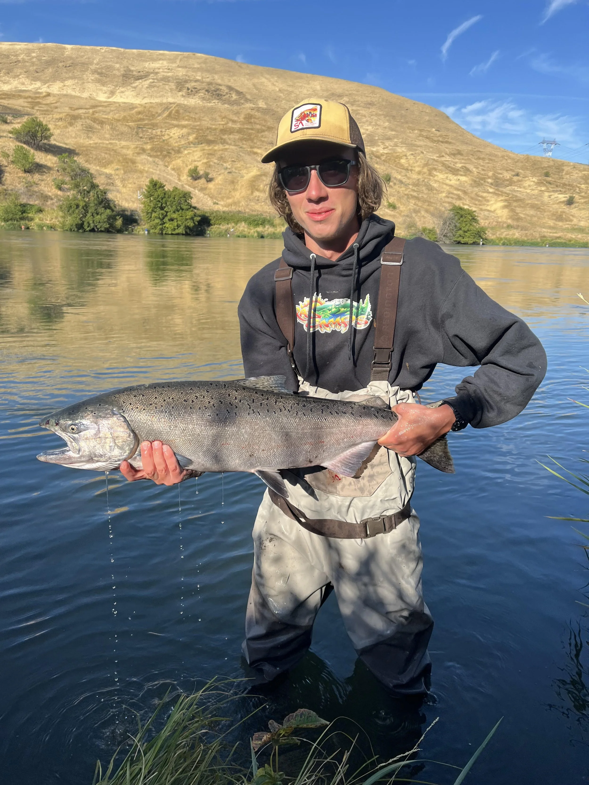 A man in fishing waders, hoodie, sunglasses, and a cap holding a large fish, standing in a river with a hillside and trees in the background on a sunny day.