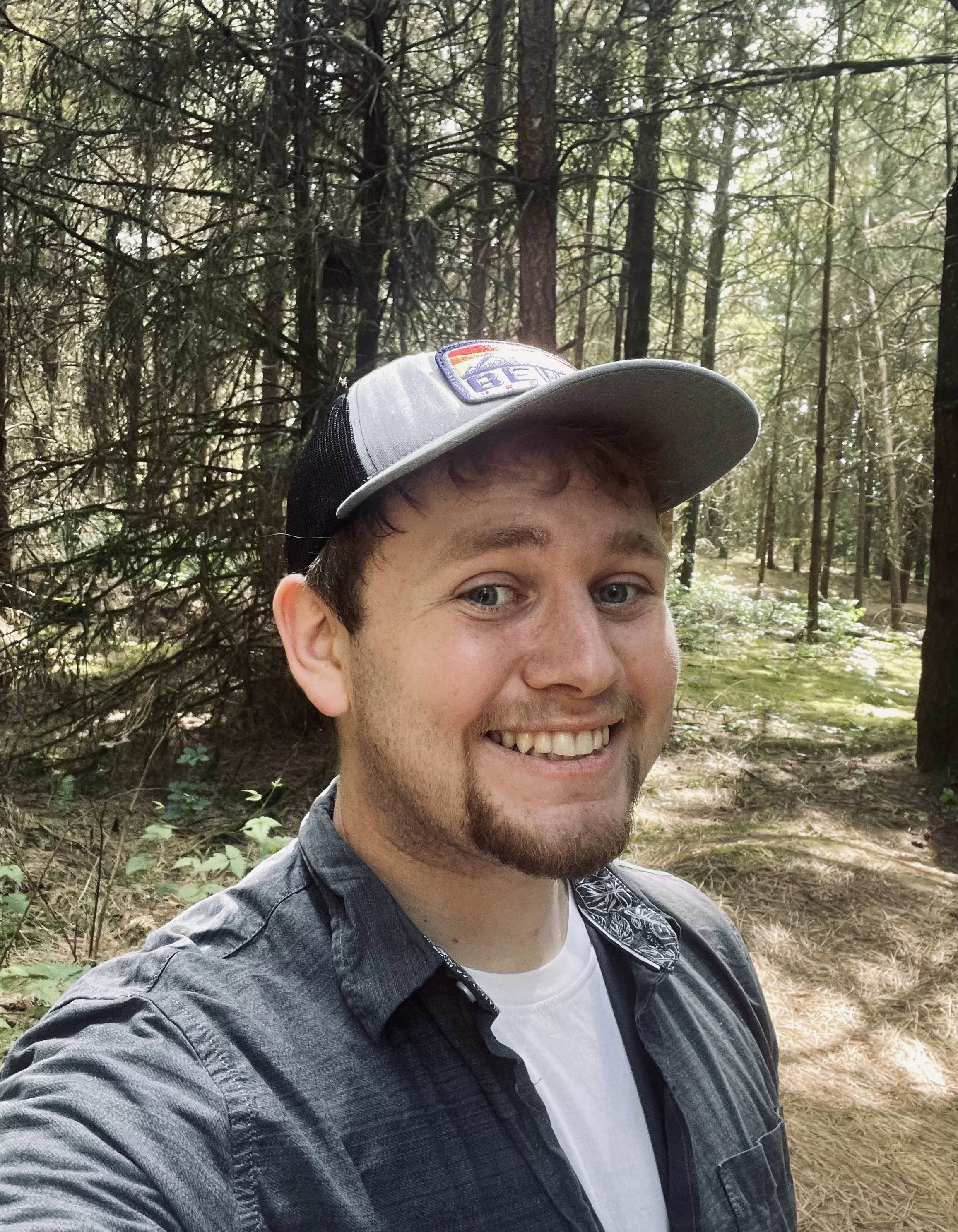 A young man smiling outdoors in a forest. He is wearing a gray baseball cap and a dark shirt over a white T-shirt.
