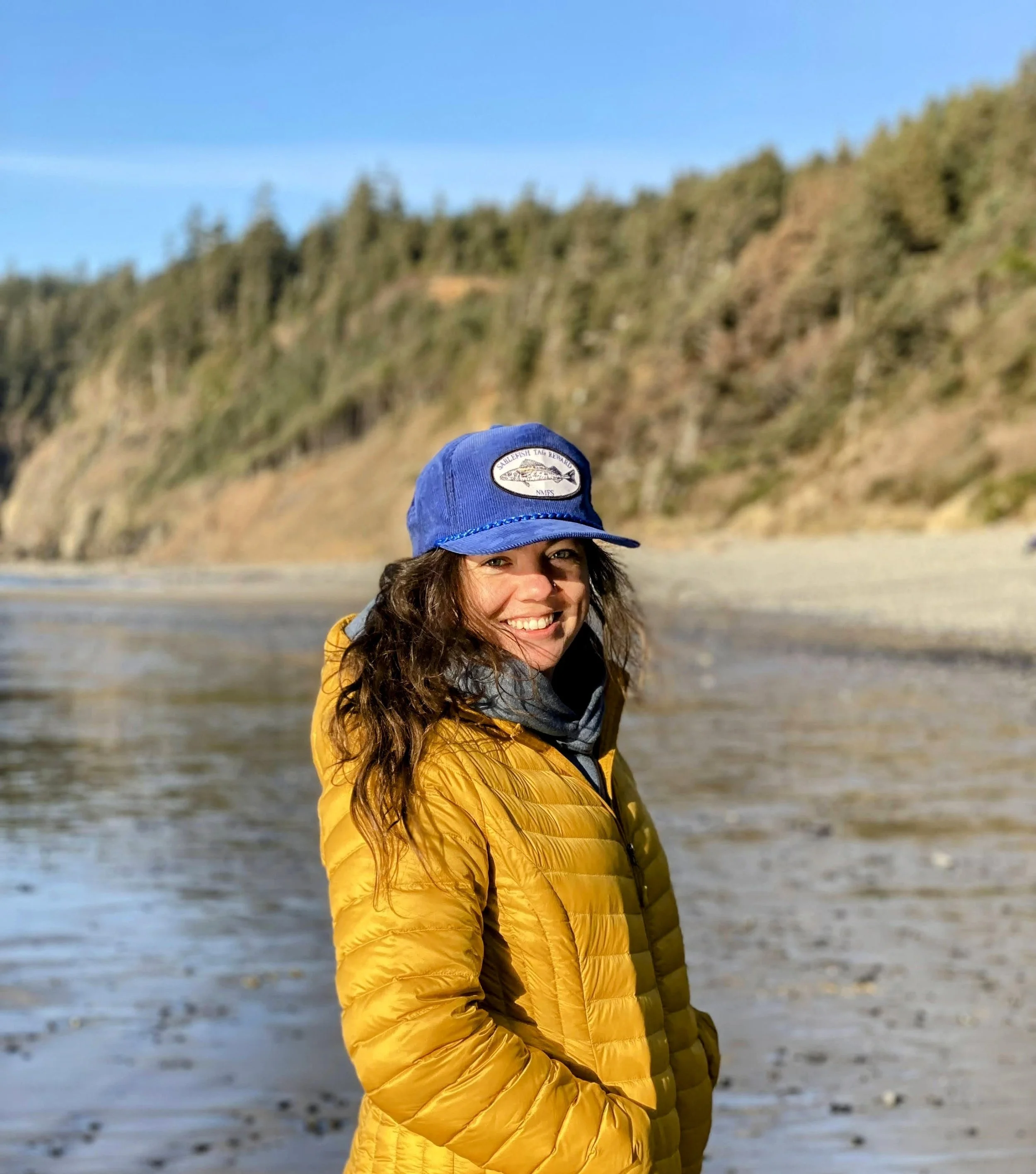 Woman smiling at the beach wearing a yellow jacket and blue cap with a mountain landscape in the background.