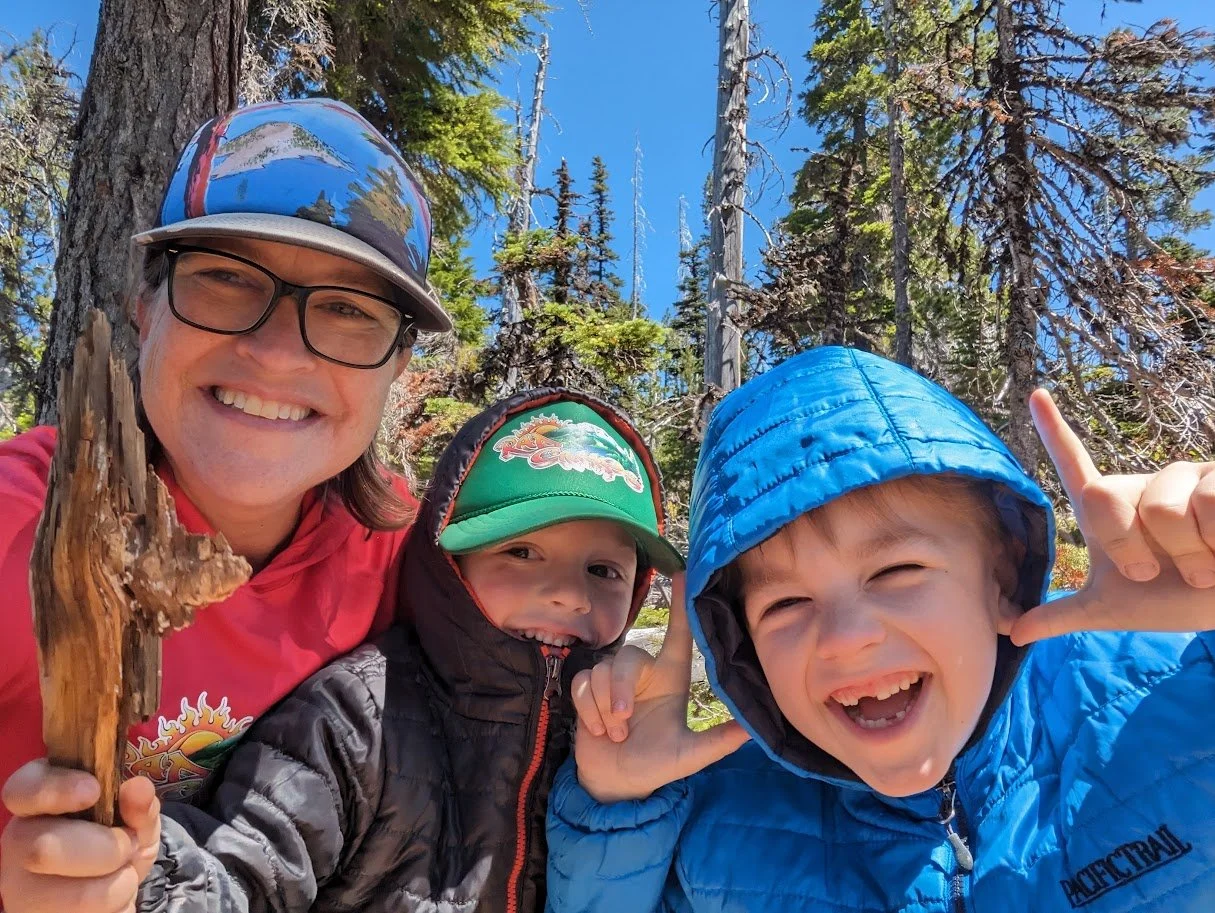 Three people smiling outdoors in a forest with tall trees, one adult woman and two young children, wearing jackets and hats, taking a selfie.