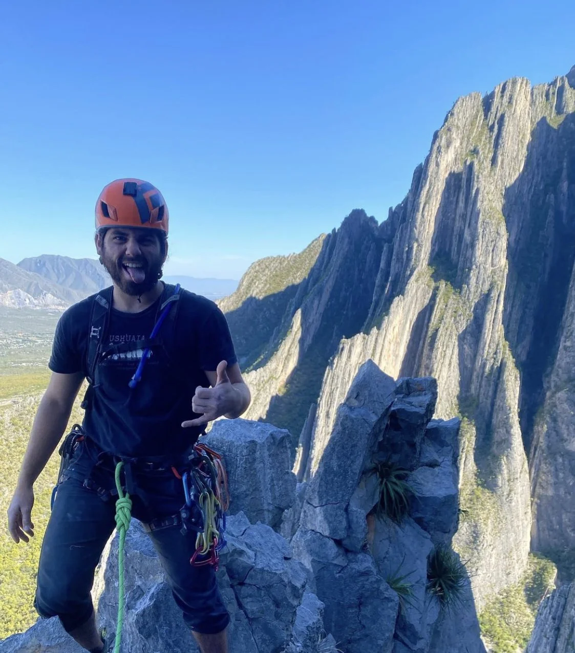 A man wearing an orange helmet and climbing gear, smiling and making a shaka sign, standing on rocks with a mountain landscape in the background.