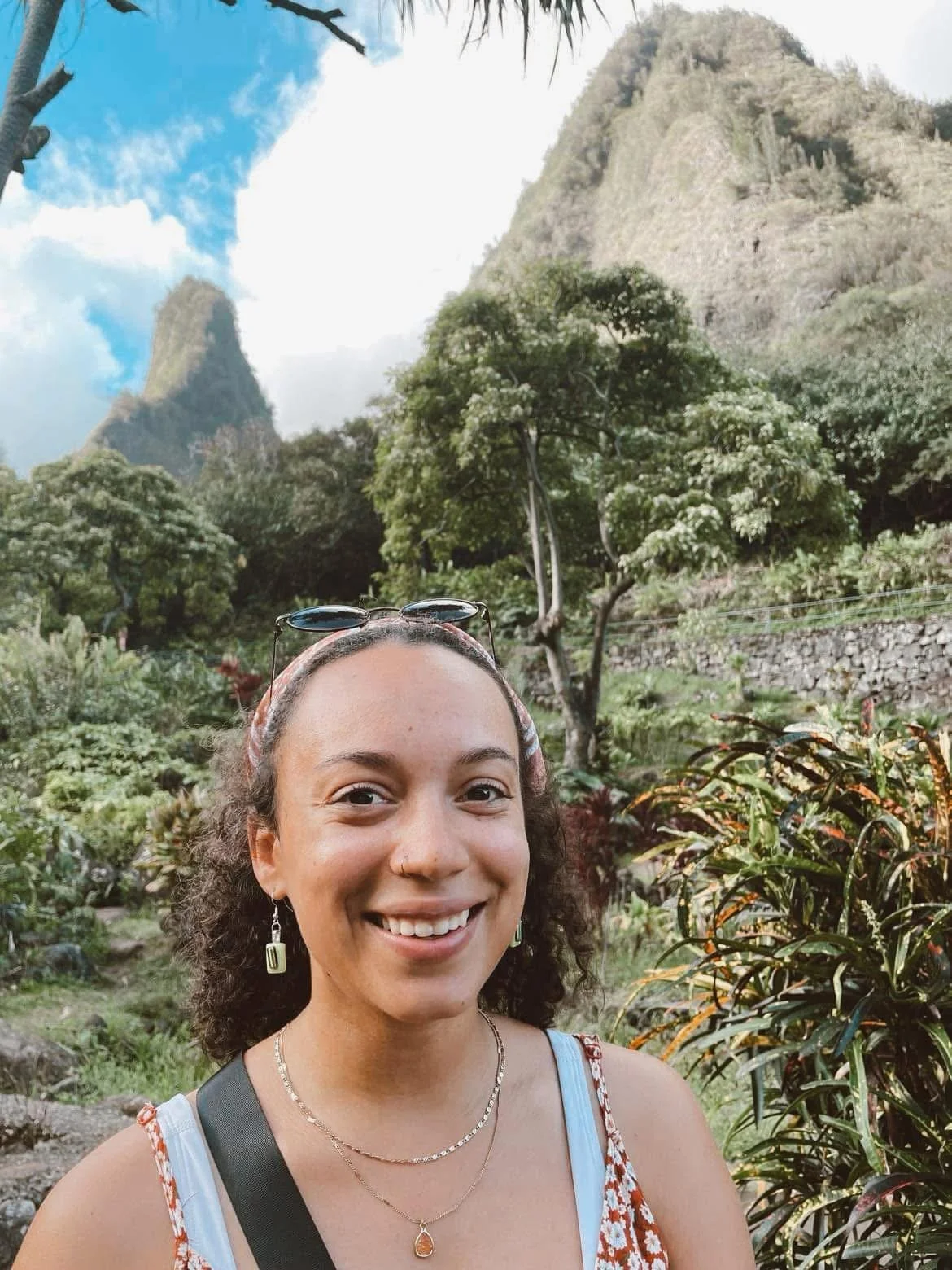 A woman smiling outdoors with lush greenery and tall mountains in the background.