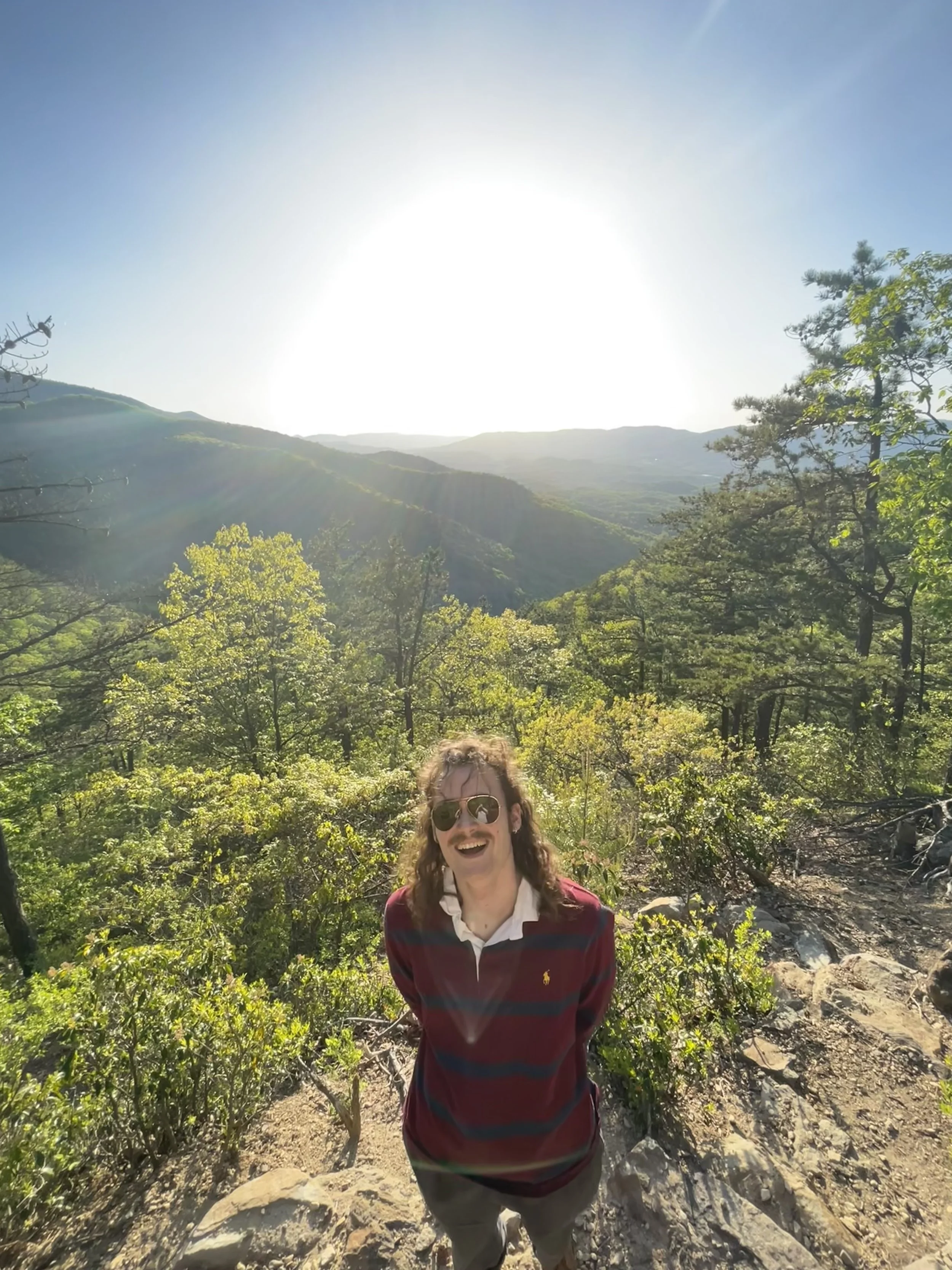 Woman wearing sunglasses smiling in a forested mountain area with the sun shining overhead.