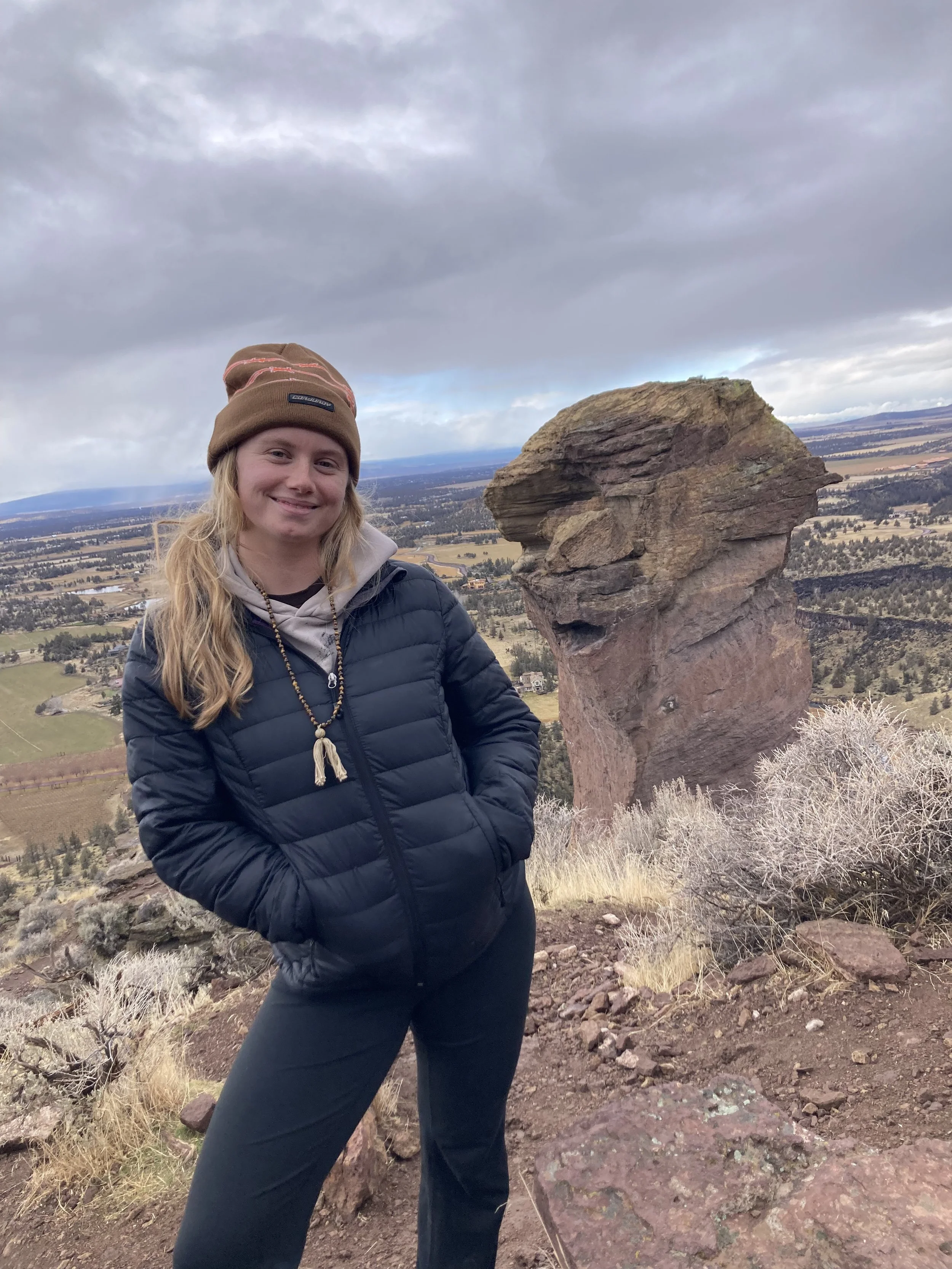A woman hiking outdoors, standing next to a large rock formation shaped like a dog, with a landscape of fields and trees in the background under a cloudy sky.