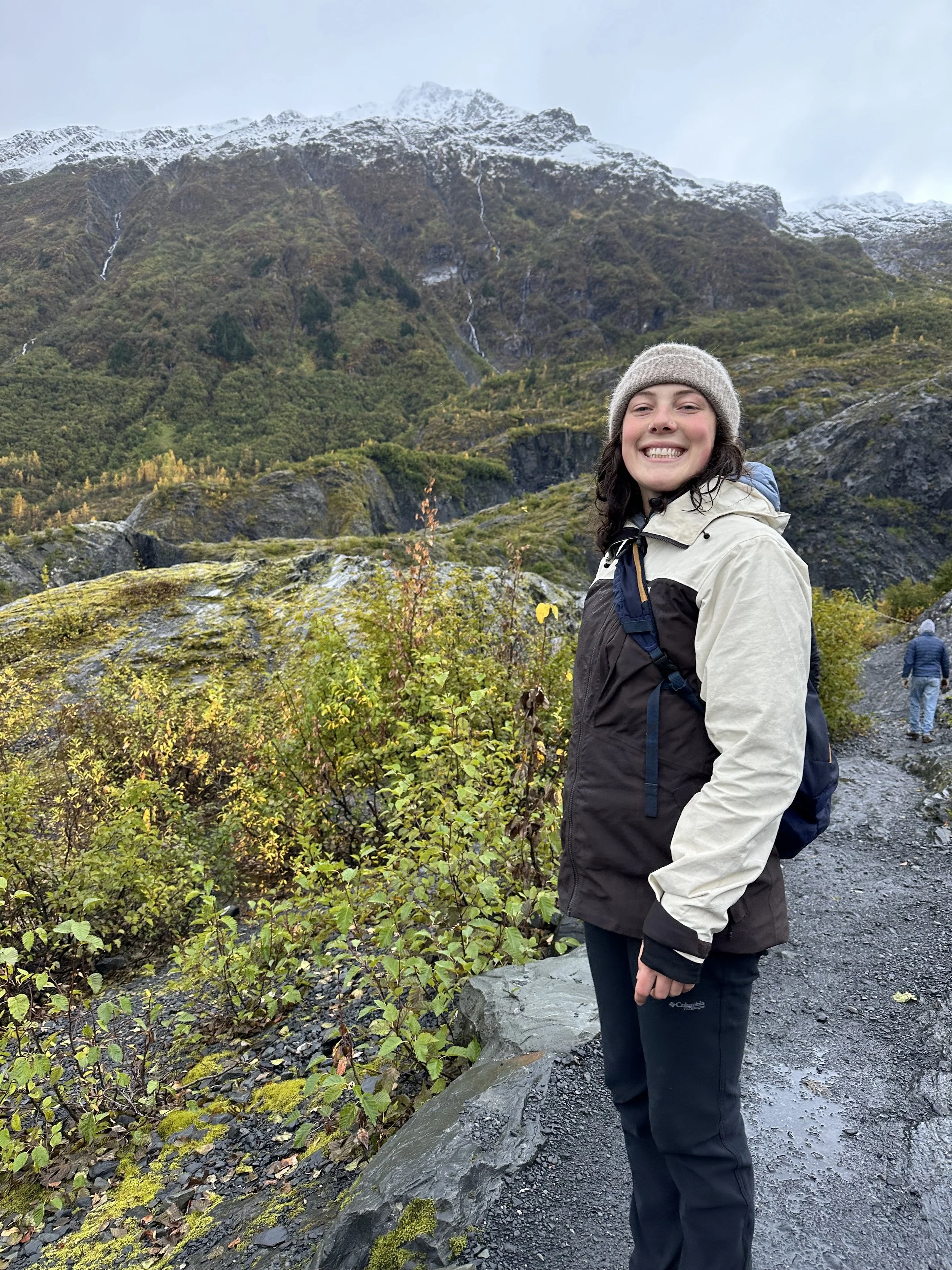 A woman with dark hair smiling in an outdoor mountainous landscape with green foliage, rocky terrain, and snow-capped peaks in the background.