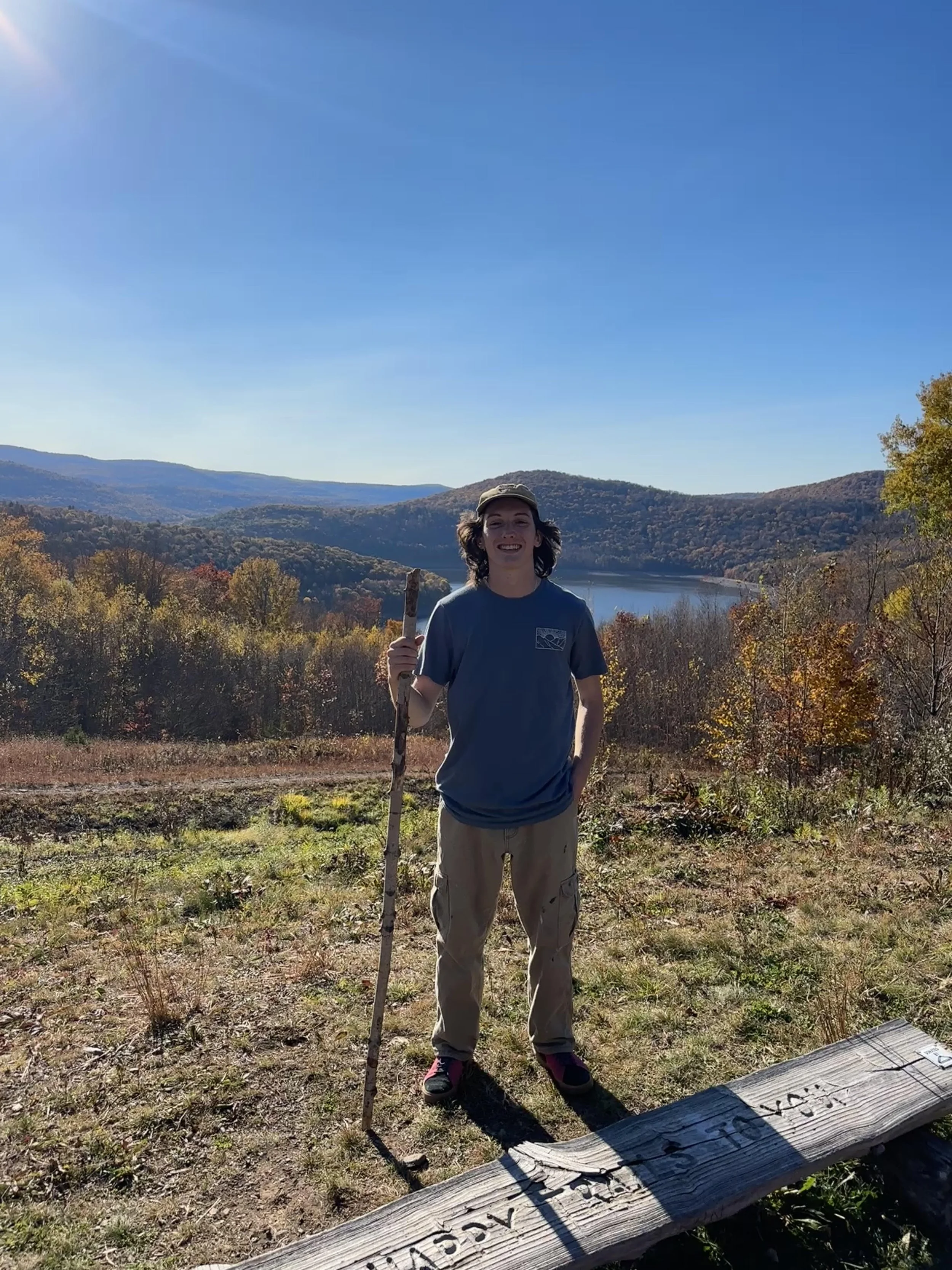 Person standing outdoors on a hilltop during autumn, holding a walking stick, with a lake and rolling hills in the background, under a clear blue sky.