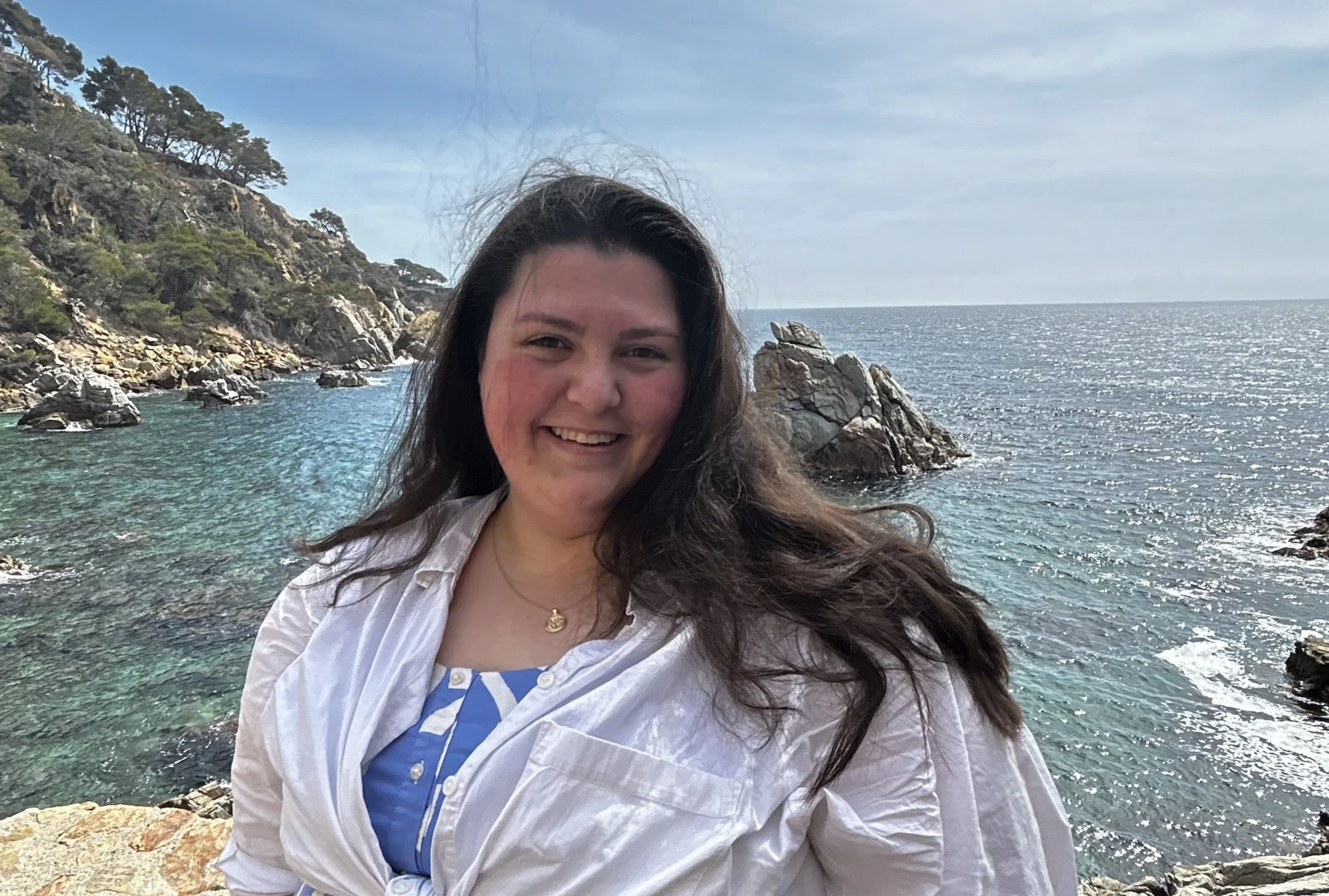 A smiling woman with long dark hair, wearing a white shirt and patterned blue dress, standing on rocky coast with the ocean and rocky cliffs in the background.