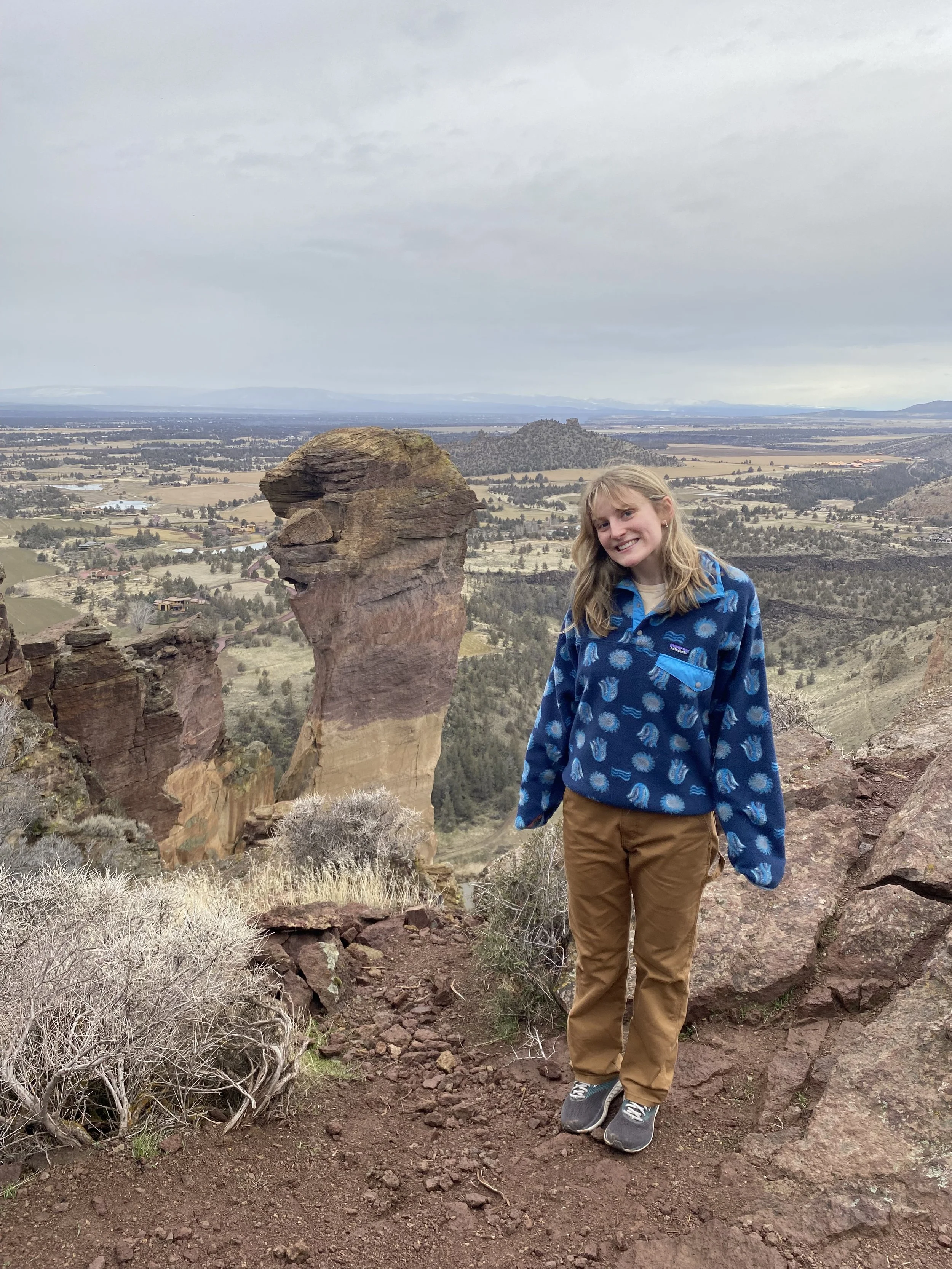A young woman standing on rocky terrain with a unique rock formation resembling a head in the background, overlooking a vast valley with rolling hills and a cloudy sky.