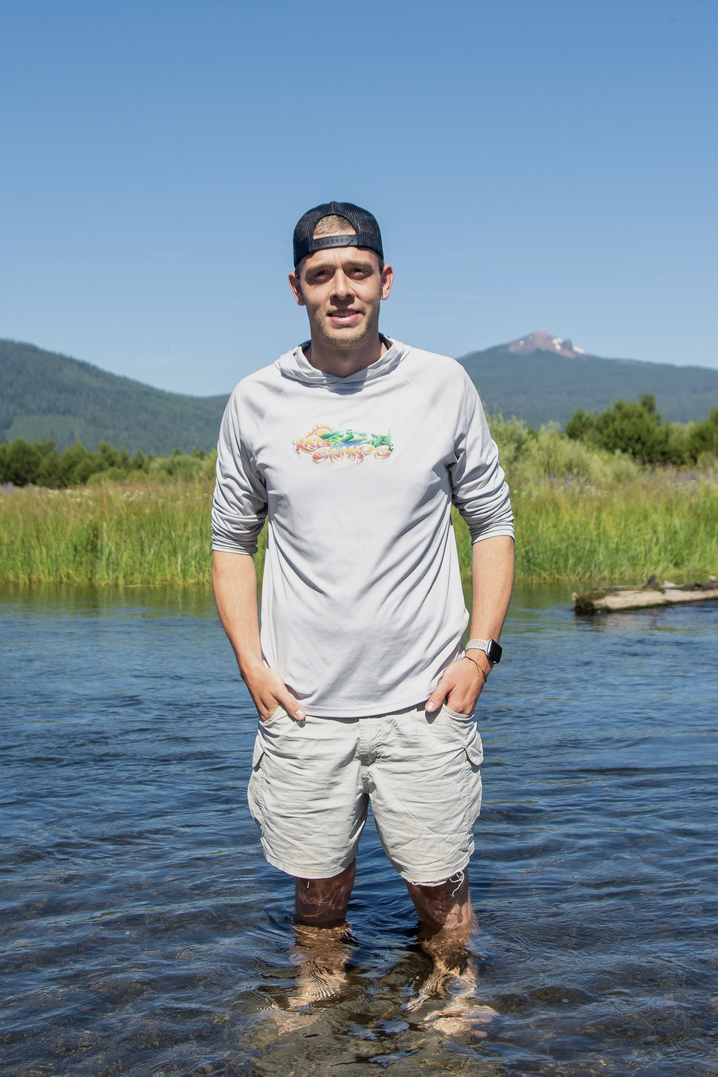 A man standing in a shallow river with green grass and mountains in the background, wearing a gray long-sleeve shirt, khaki shorts, a black cap backwards, and a smartwatch.