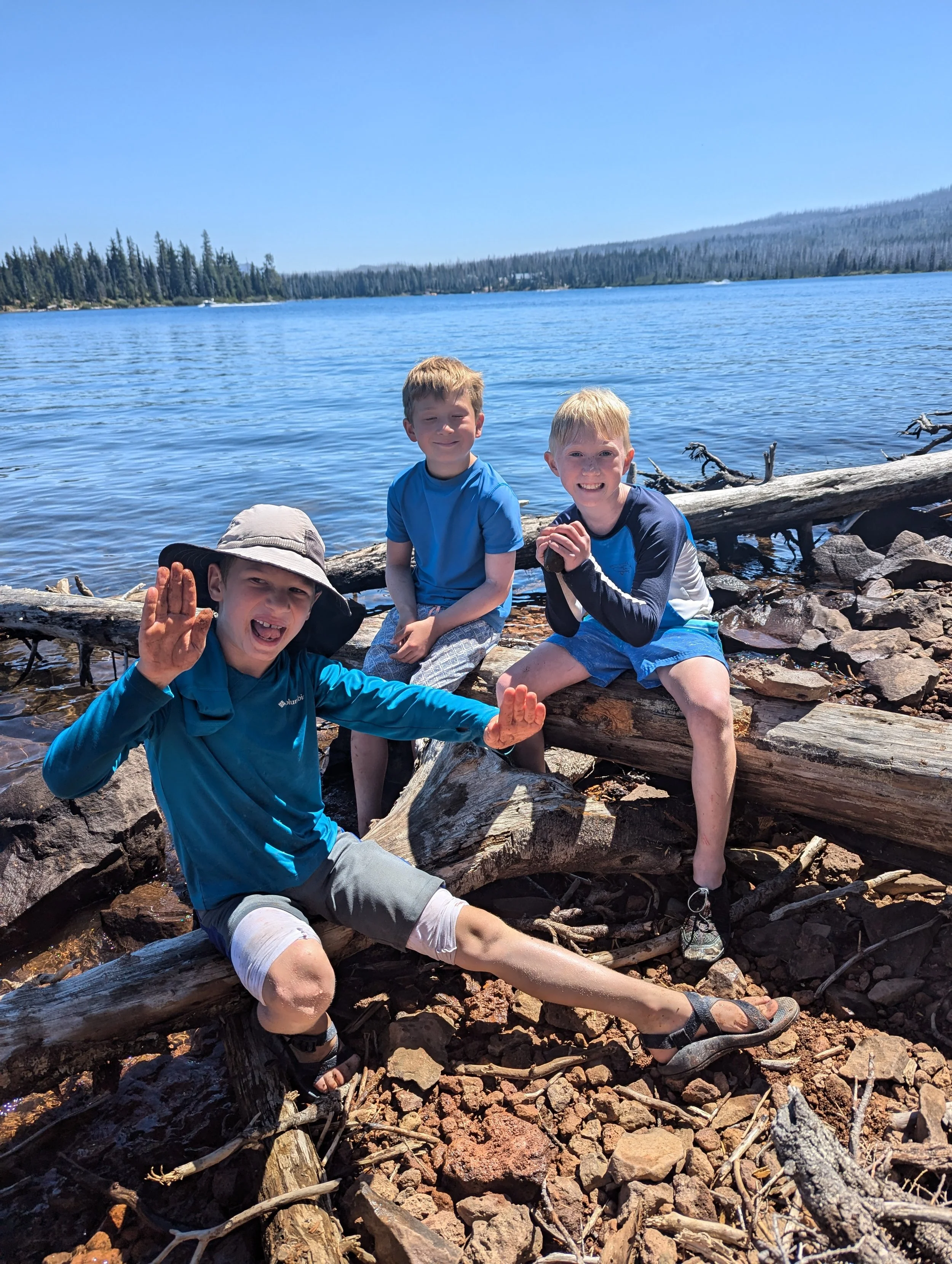 Three boys by a lake with trees and a mountain in the background, sitting on logs and rocks, smiling.