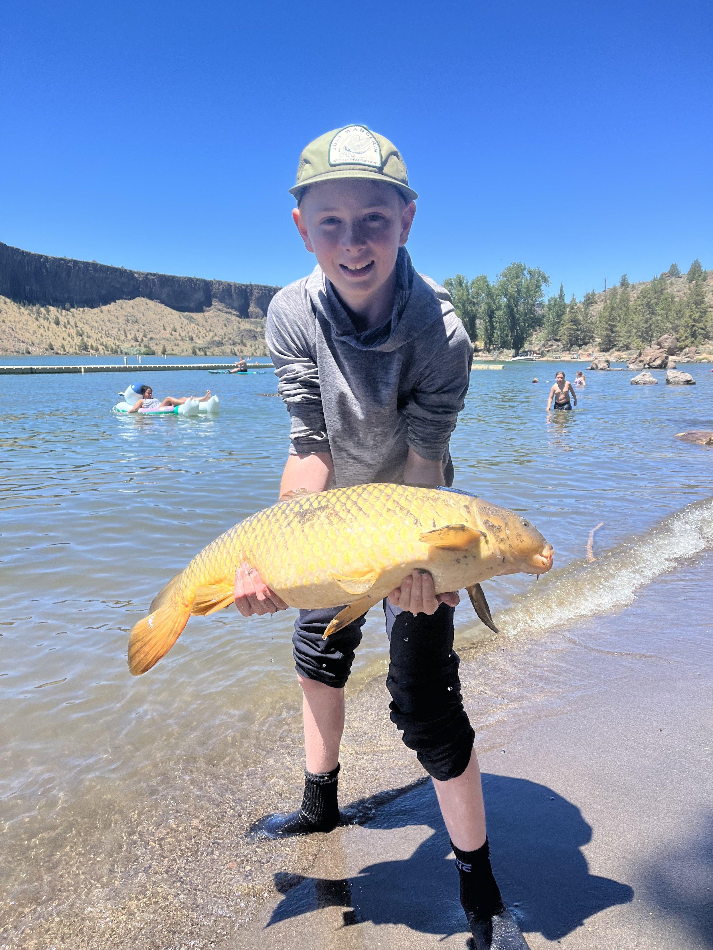Young boy smiling and holding a large yellow carp fish at a lake, with other swimmers and a boat in the background under a clear blue sky.