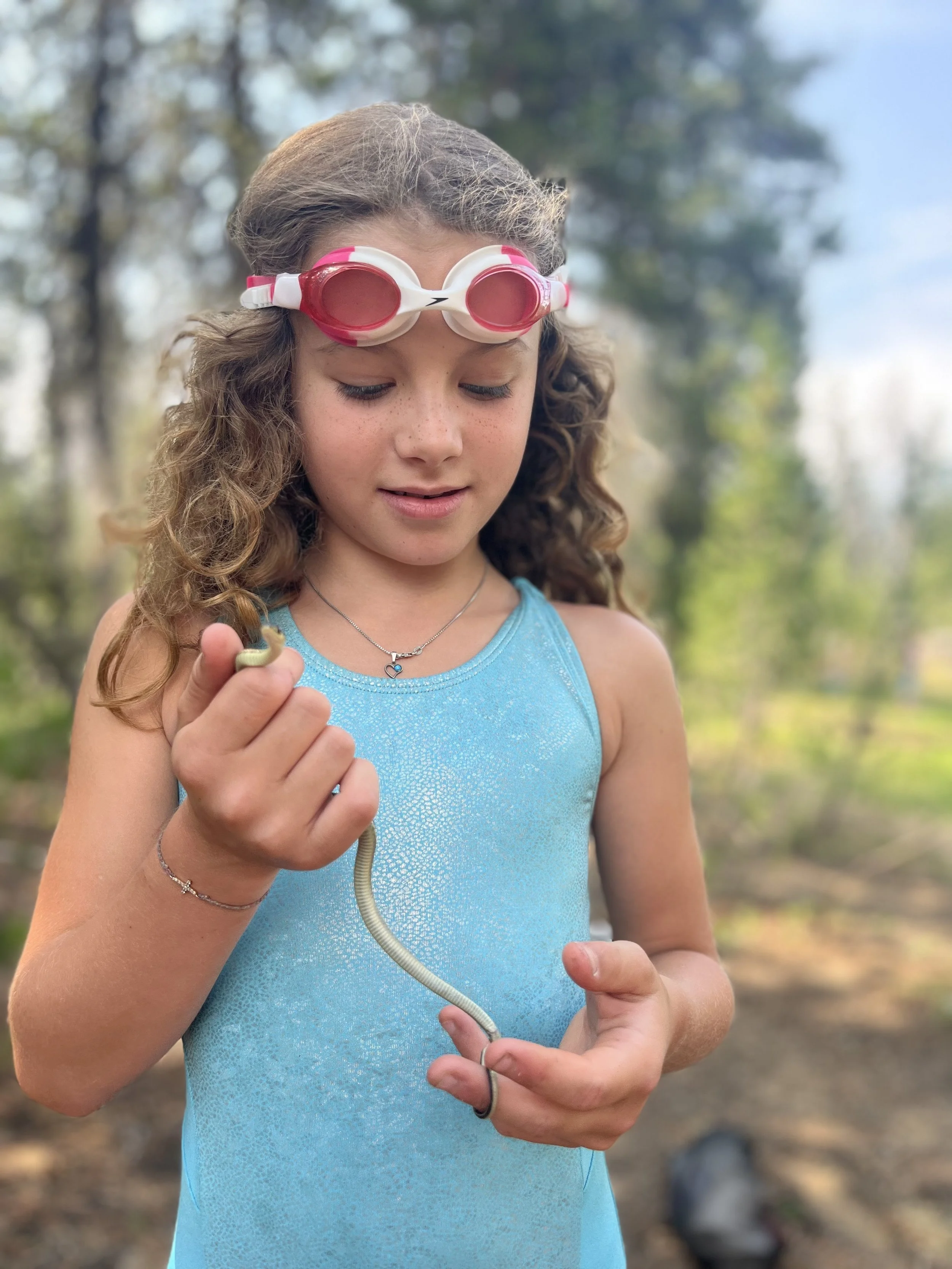 Young girl with goggles holding a small snake outdoors in a wooded area.