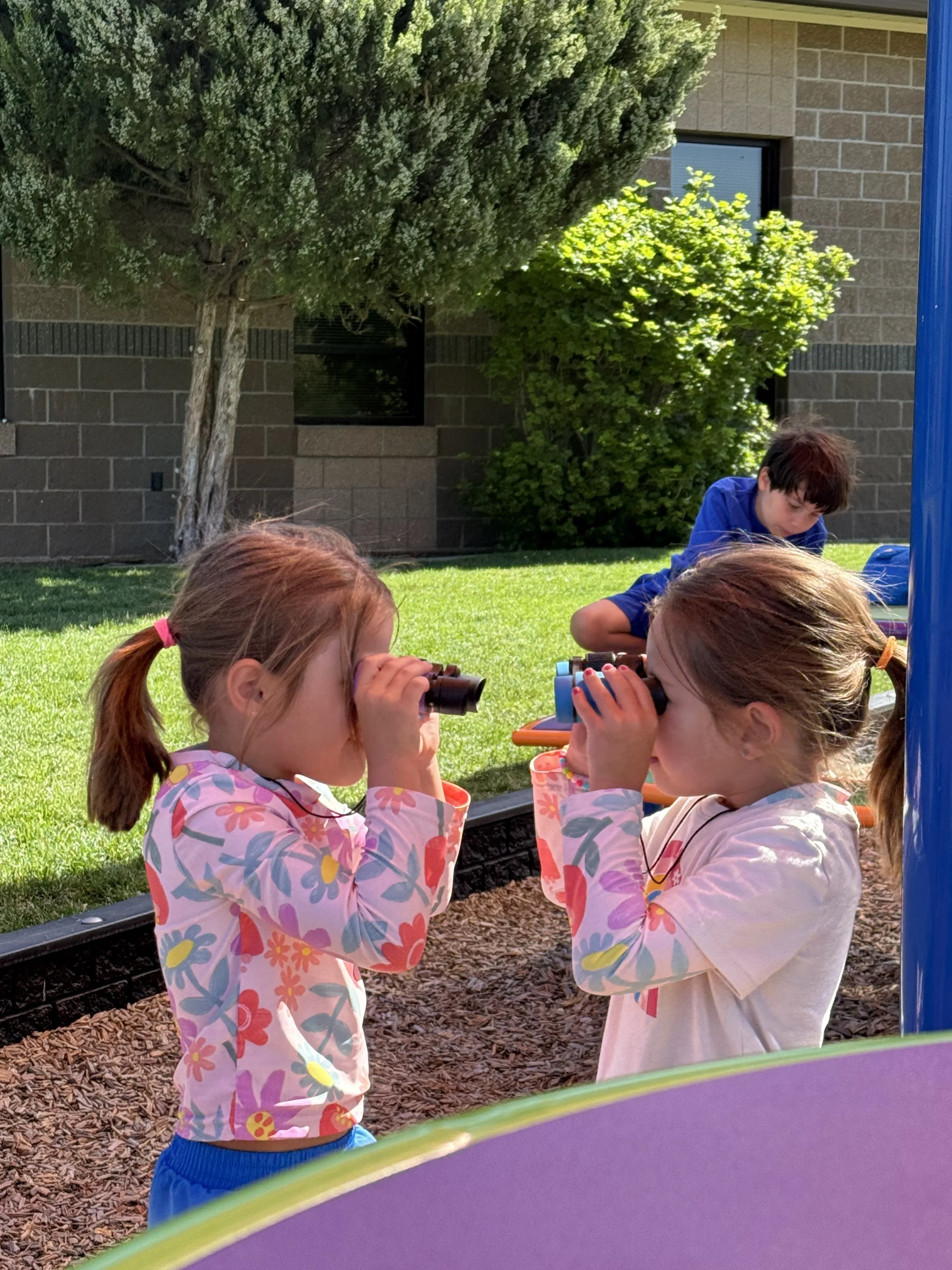Two young girls standing face-to-face, looking through small binoculars, while a young boy sits on the grass in the background, outdoors near trees and a building.