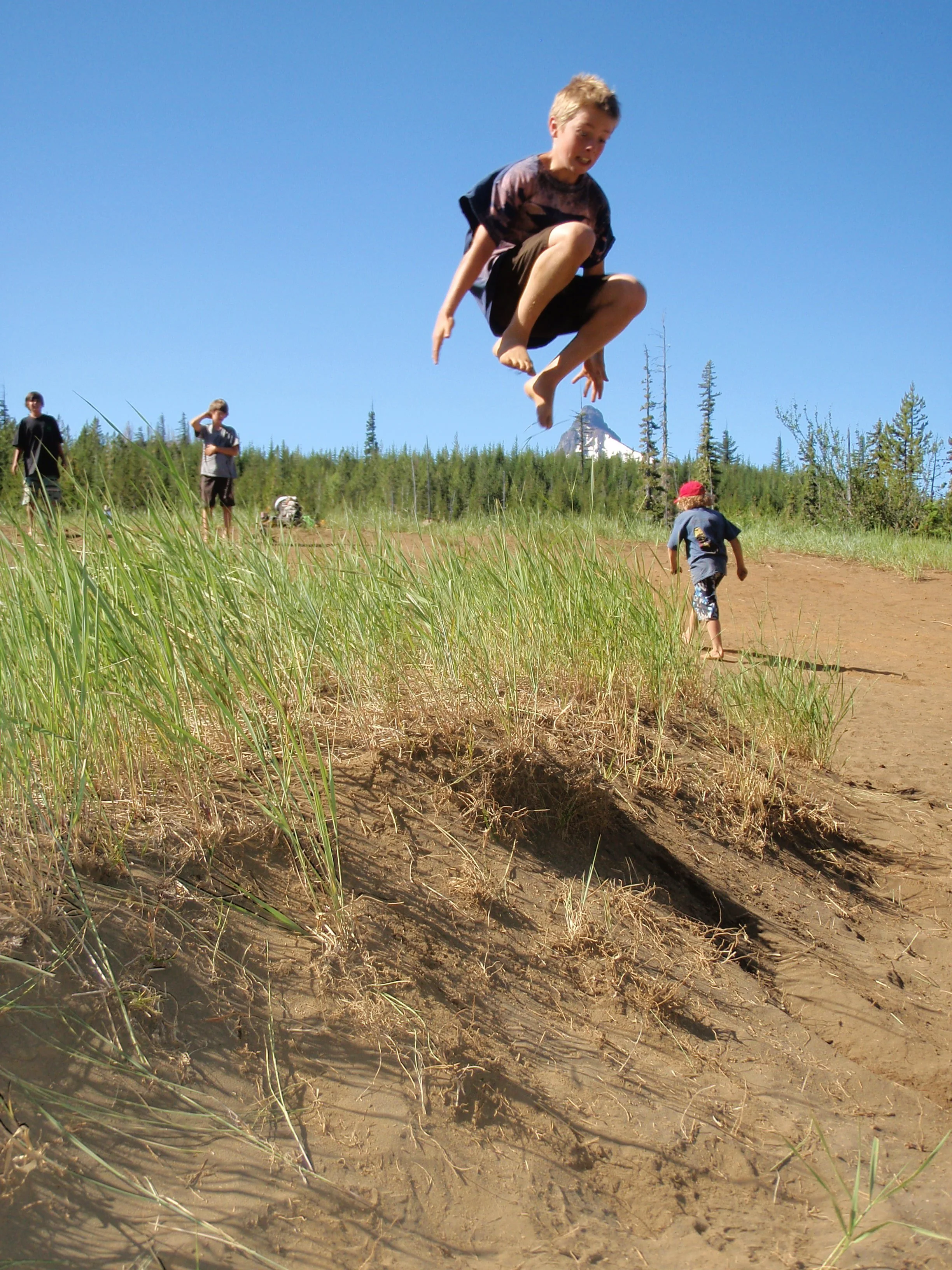 A boy mid-air jumping over a dirt trail in a grassy field with other children and adults in the background, with trees and blue sky.