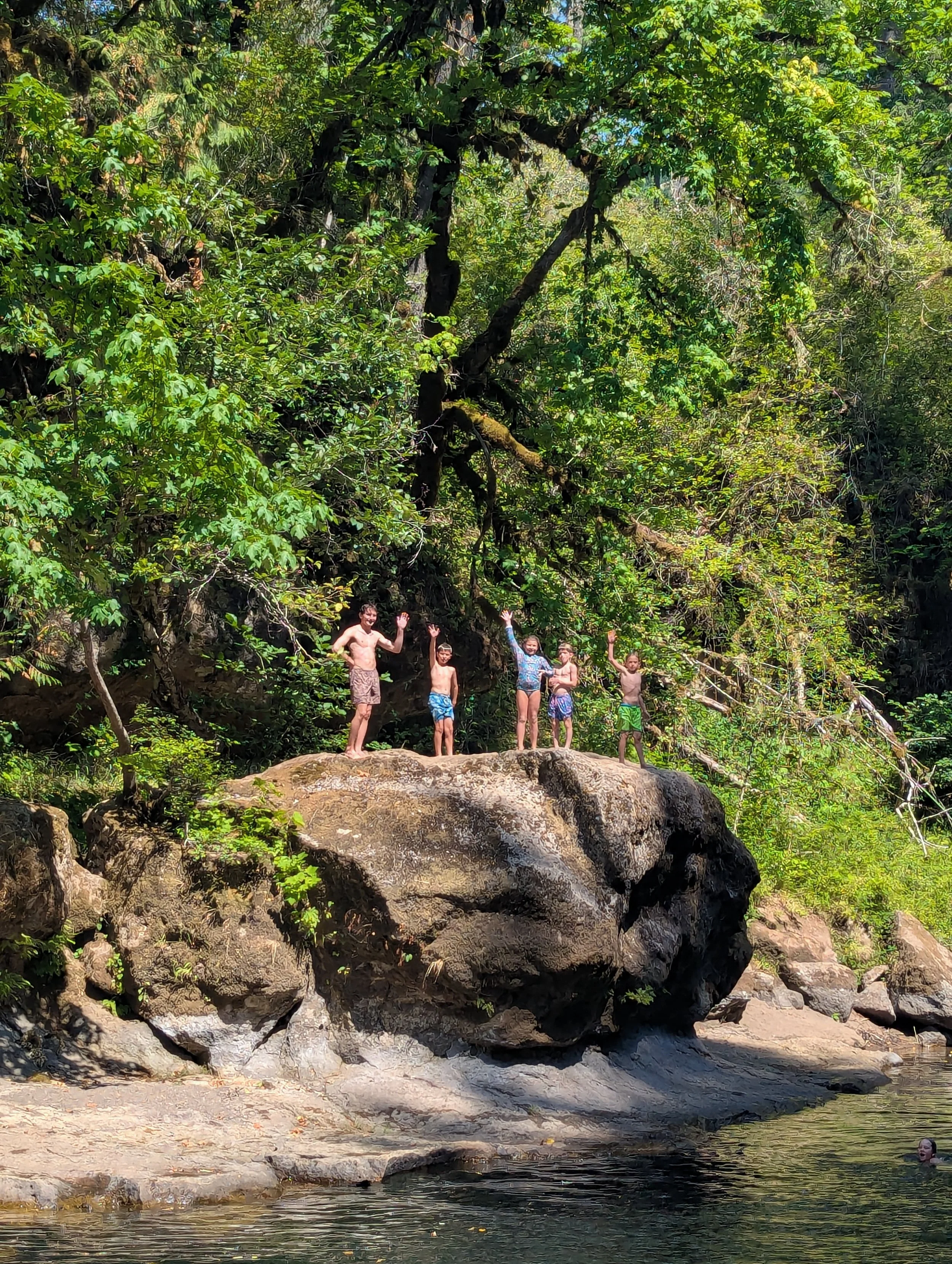 Group of children standing on a large rock by a river, surrounded by dense green forest, waving and playing in a sunny outdoor setting.
