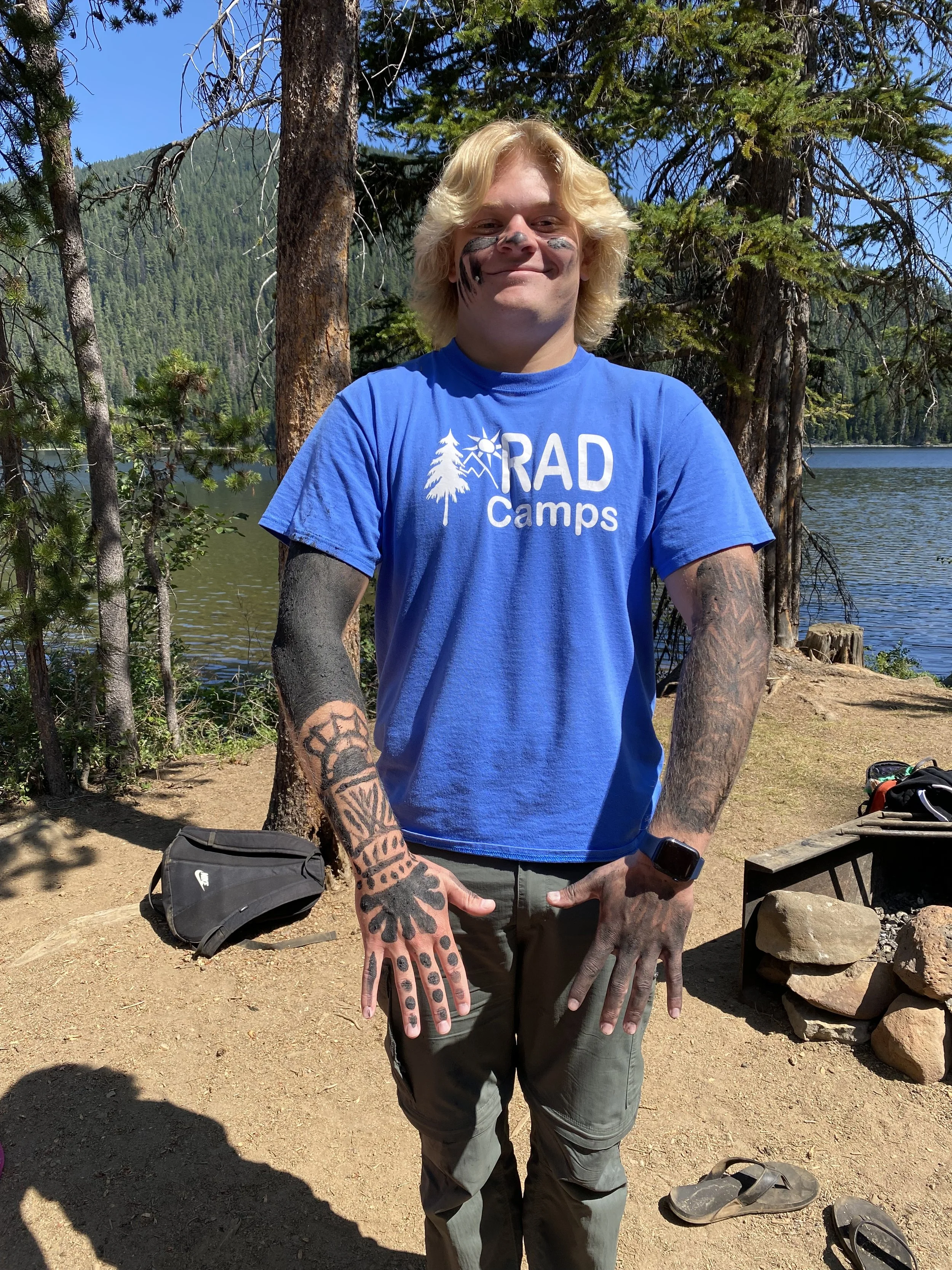 A young man with blond hair, face paint, and tattoos on his arms is standing outdoors near a lake and trees, wearing a blue 'RAD Camps' t-shirt, with a watch on his wrist, and smiling at the camera.