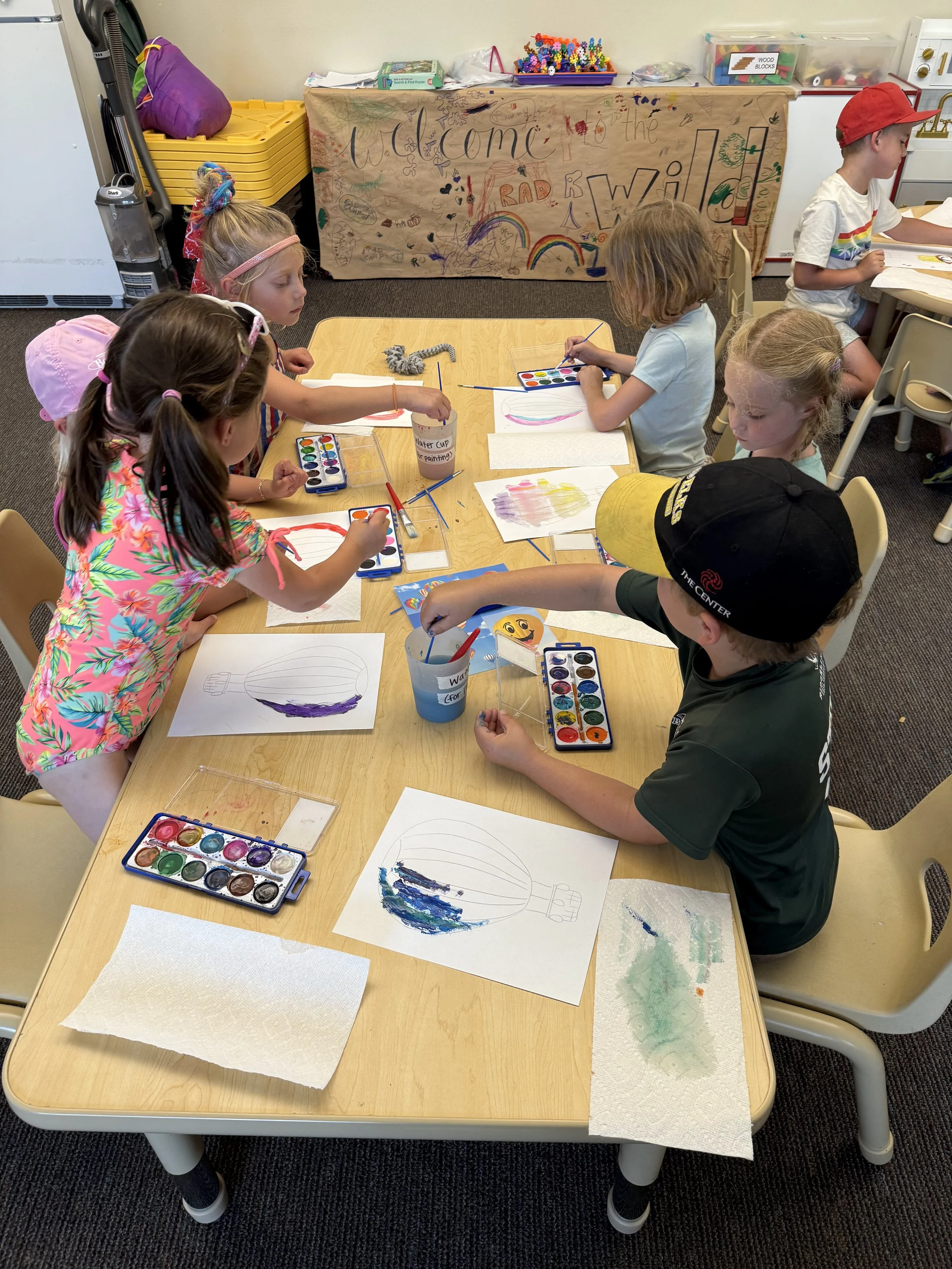 Children painting and drawing at a table in a classroom. Artwork features balloons and rainbow colors. Classroom decorations include a colorful banner and art supplies.