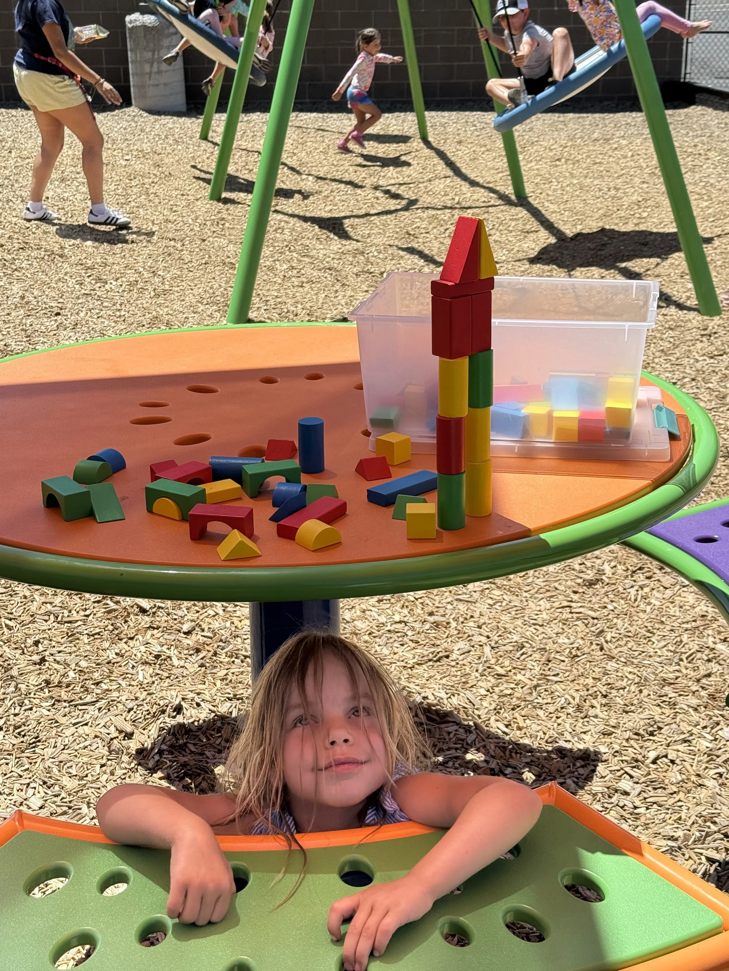 A young girl in a sandbox area of a playground, leaning over the edge of a green and orange table with large holes in it, looking upward. Behind her, there is a round orange table with colorful building blocks stacked into a tower and scattered block